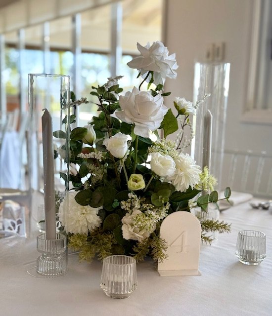 Elegant table centerpiece with white flowers in a glass vase, surrounded by tall and short candles in glass holders, on a white table in a decorated indoor space.