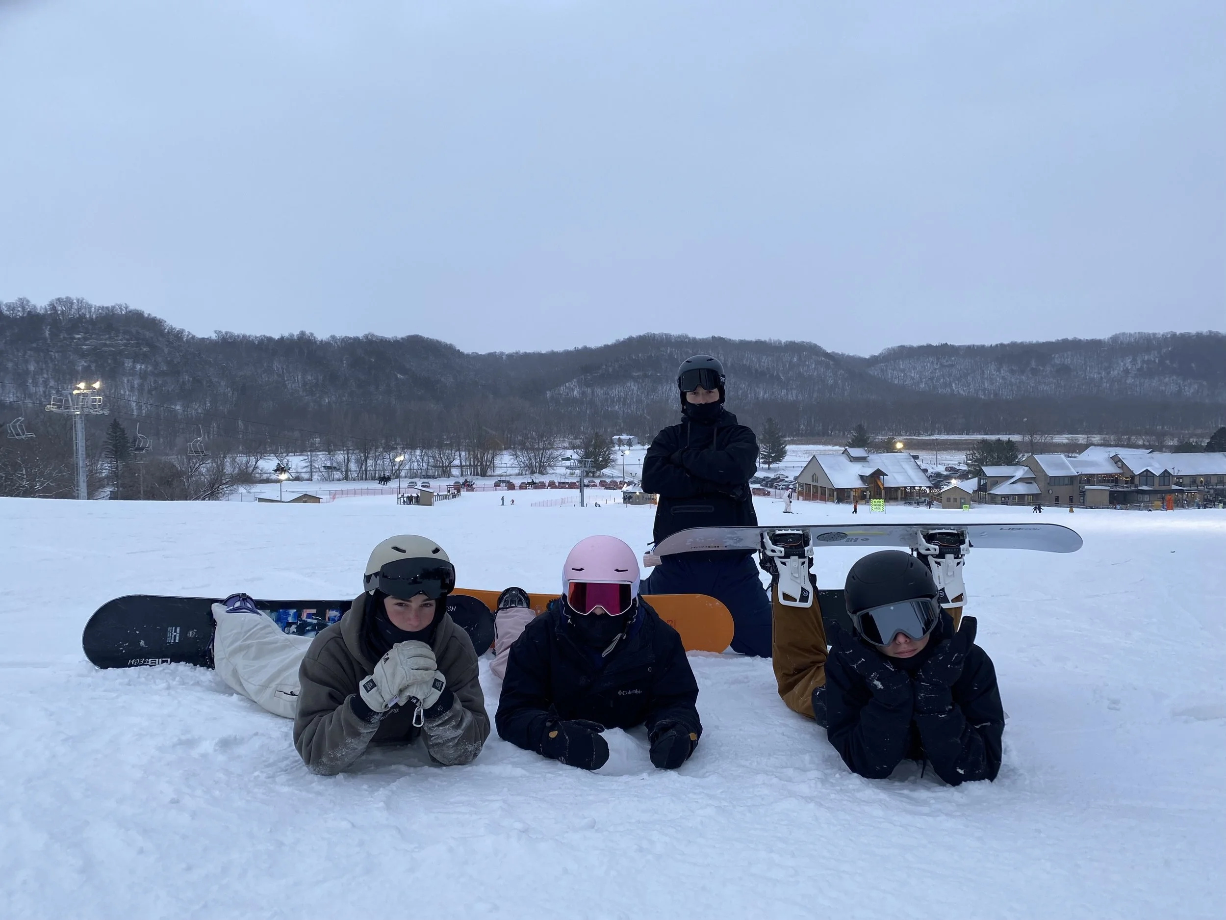Four people in winter gear and helmets on a snowy ski slope, with three lying in the snow and one standing behind their ski equipment, mountains and ski resort buildings in the background.