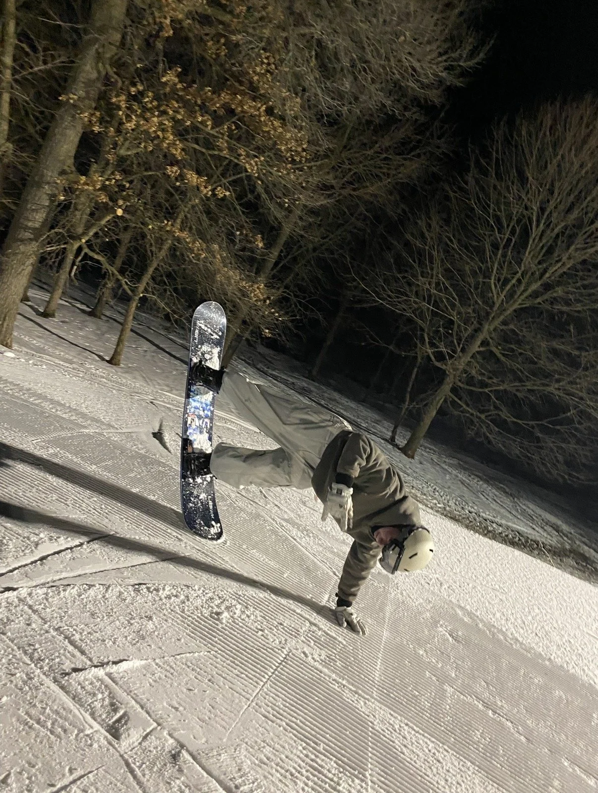 Person in winter gear balancing on one hand on snowy ground with snowboard nearby, holding a hand to support themselves, at night with leafless trees in the background.