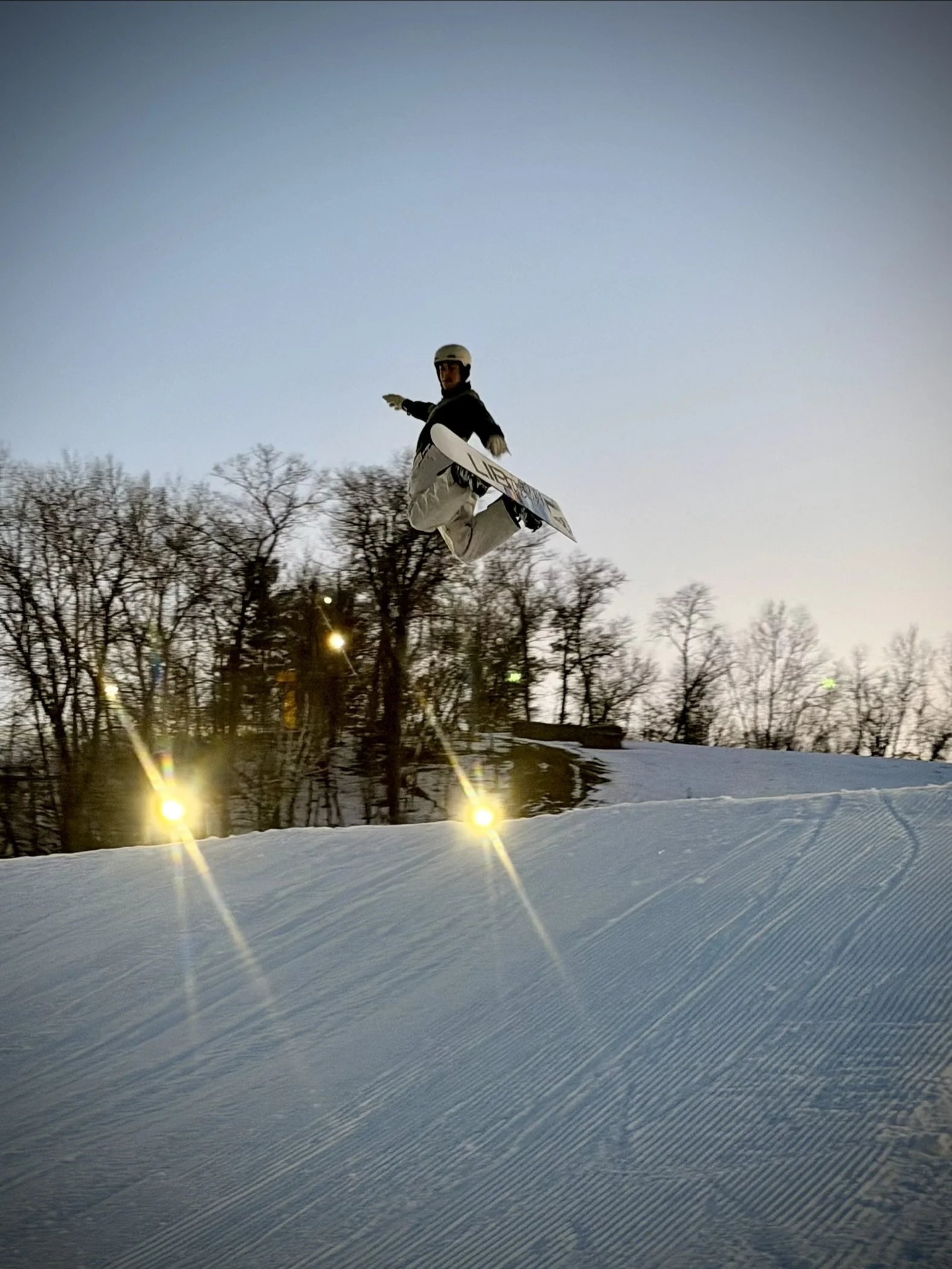 Skier airborne on a snow-covered slope during dusk or dawn, with leafless trees and bright lights in the background.