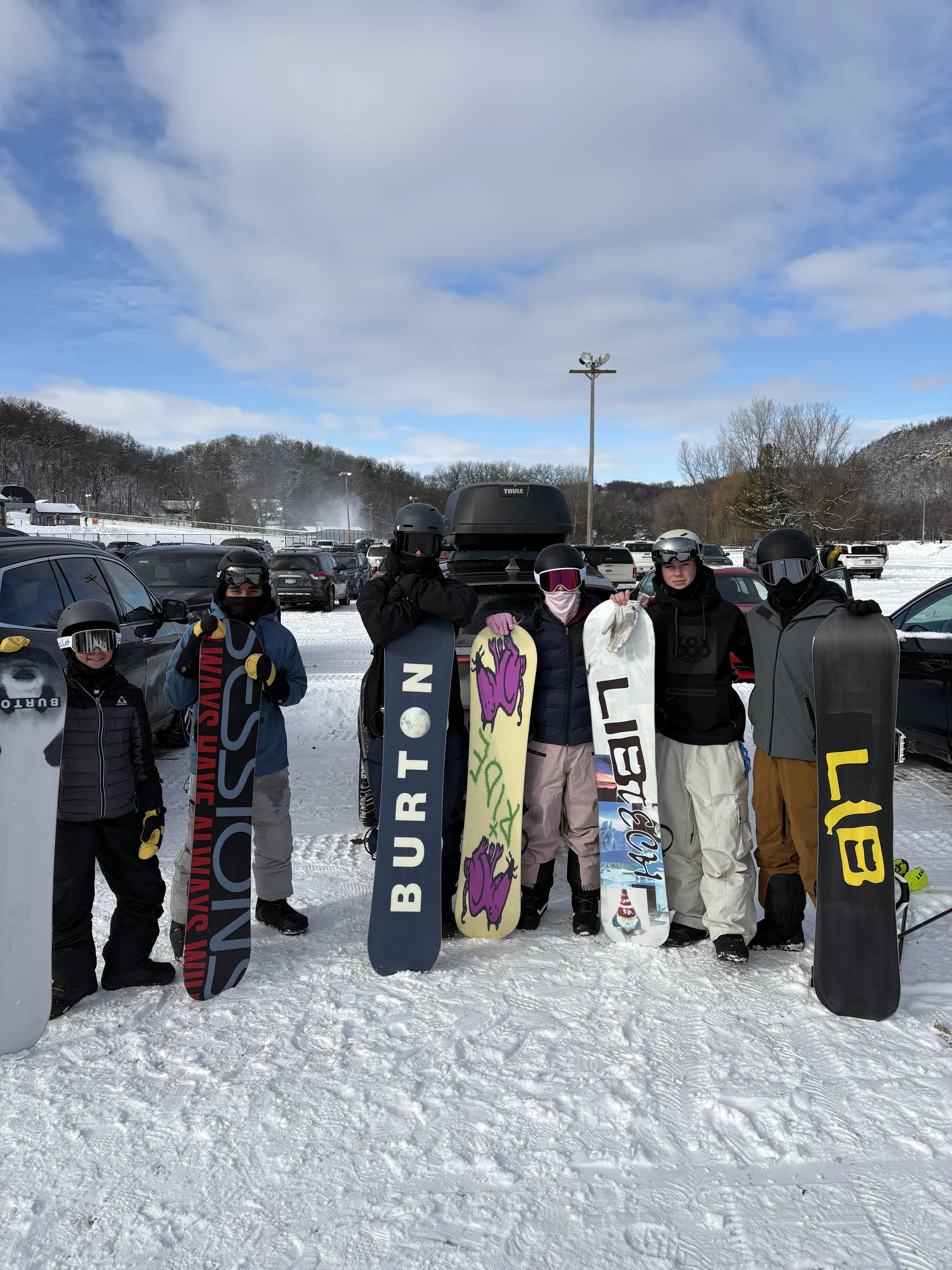 Group of six snowboarders in winter gear standing in snow, holding snowboards, with a snowy parking lot and mountains in the background.