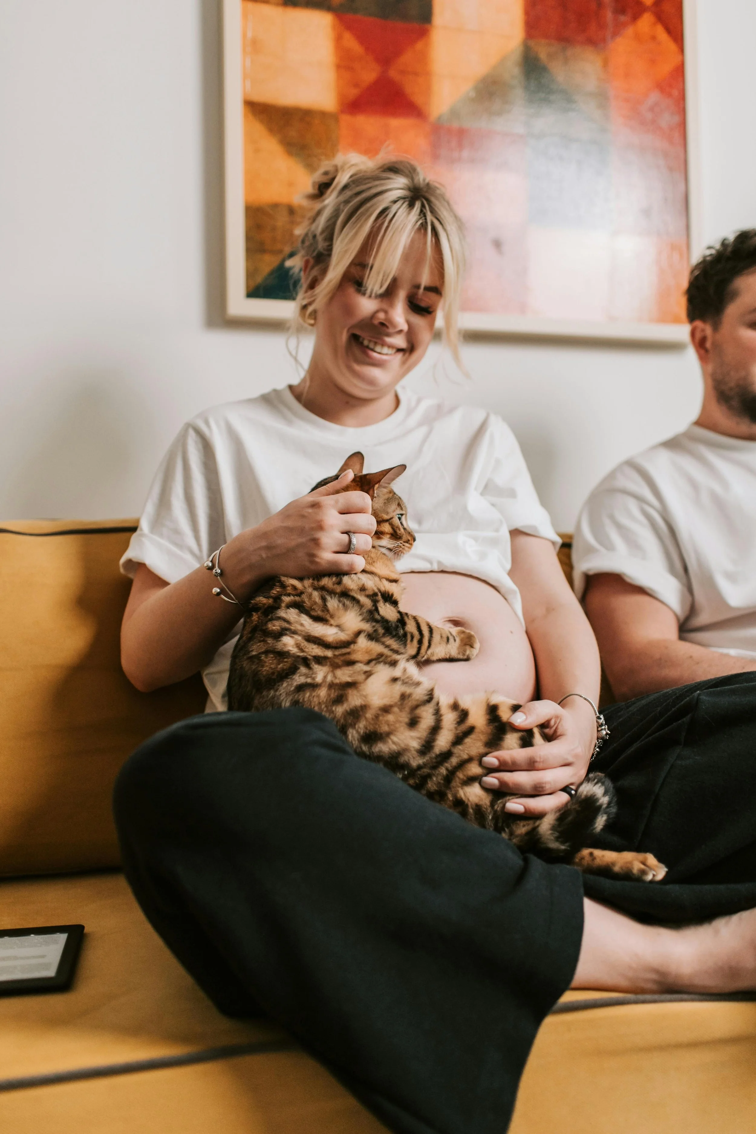 A pregnant, smiling woman with blonde hair sitting on a couch, holding a Bengal cat.