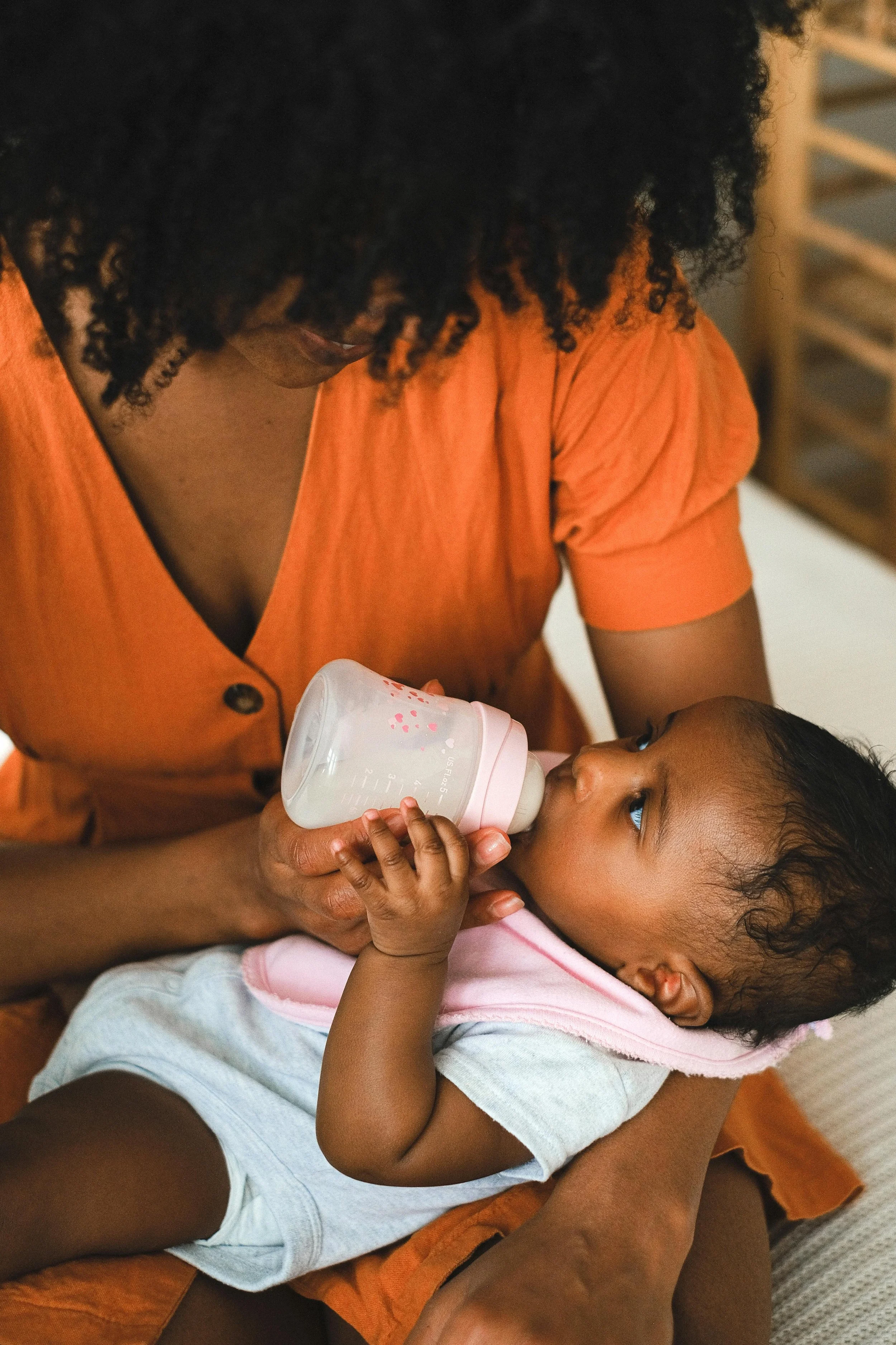 A woman in an orange top feeding a baby with a baby bottle.