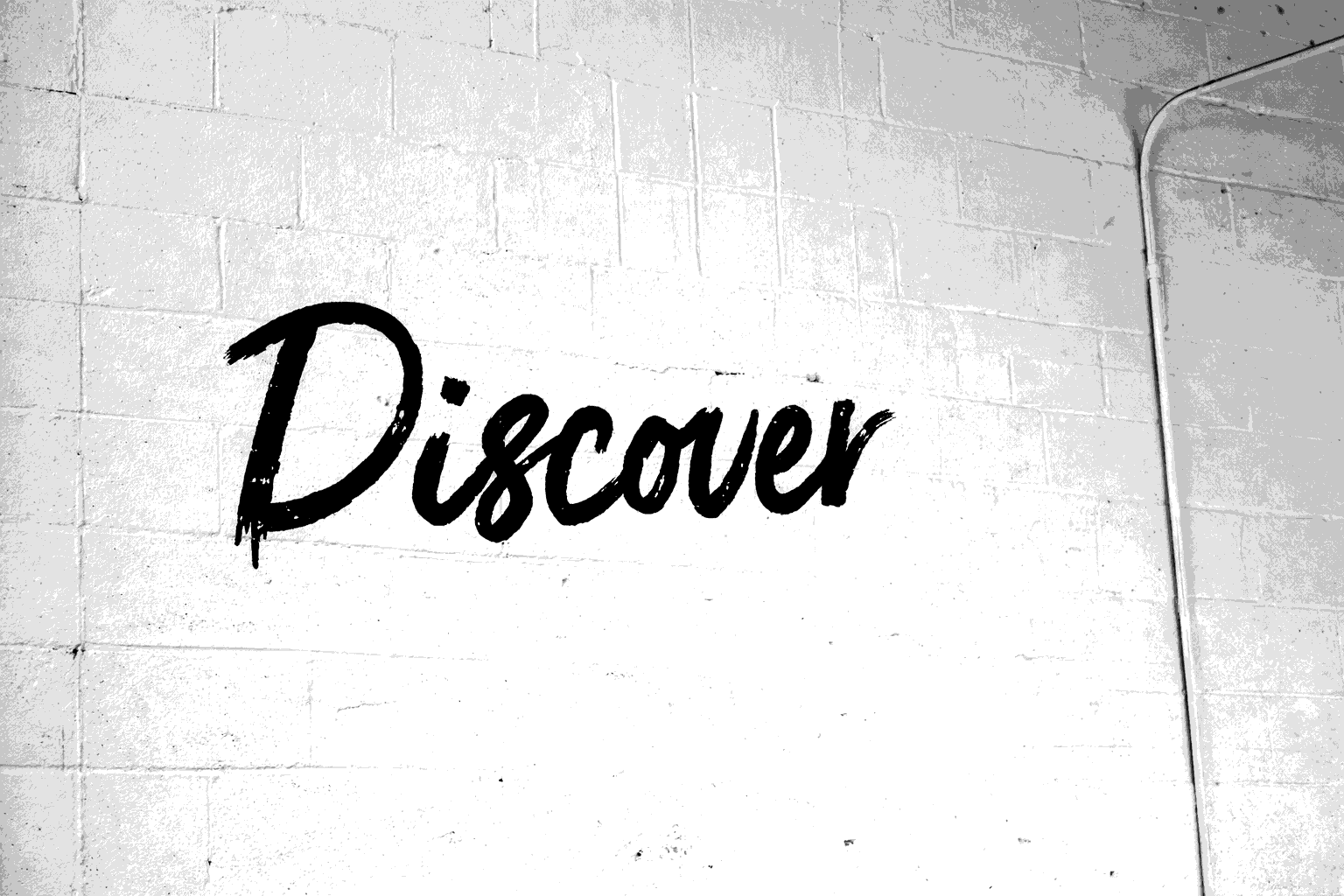 Black graffiti-style word 'Discover' on a white painted brick wall with visible piping in the upper right corner.