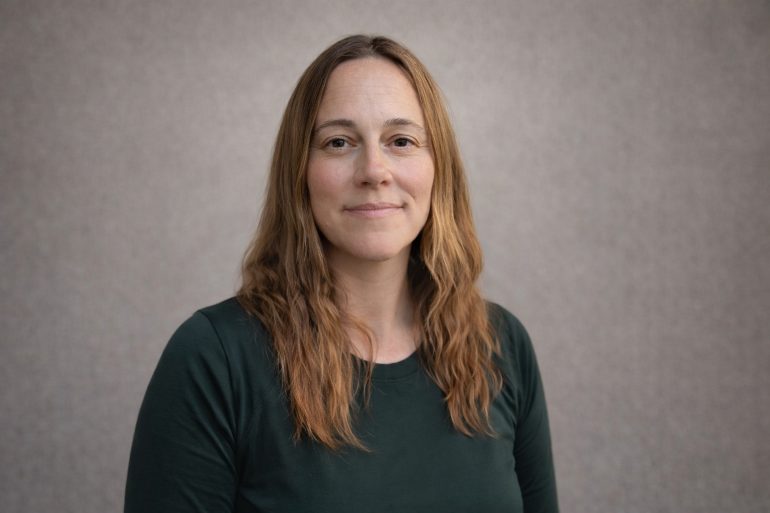 A woman with shoulder-length wavy red hair, wearing a dark green shirt, standing against a plain gray background.