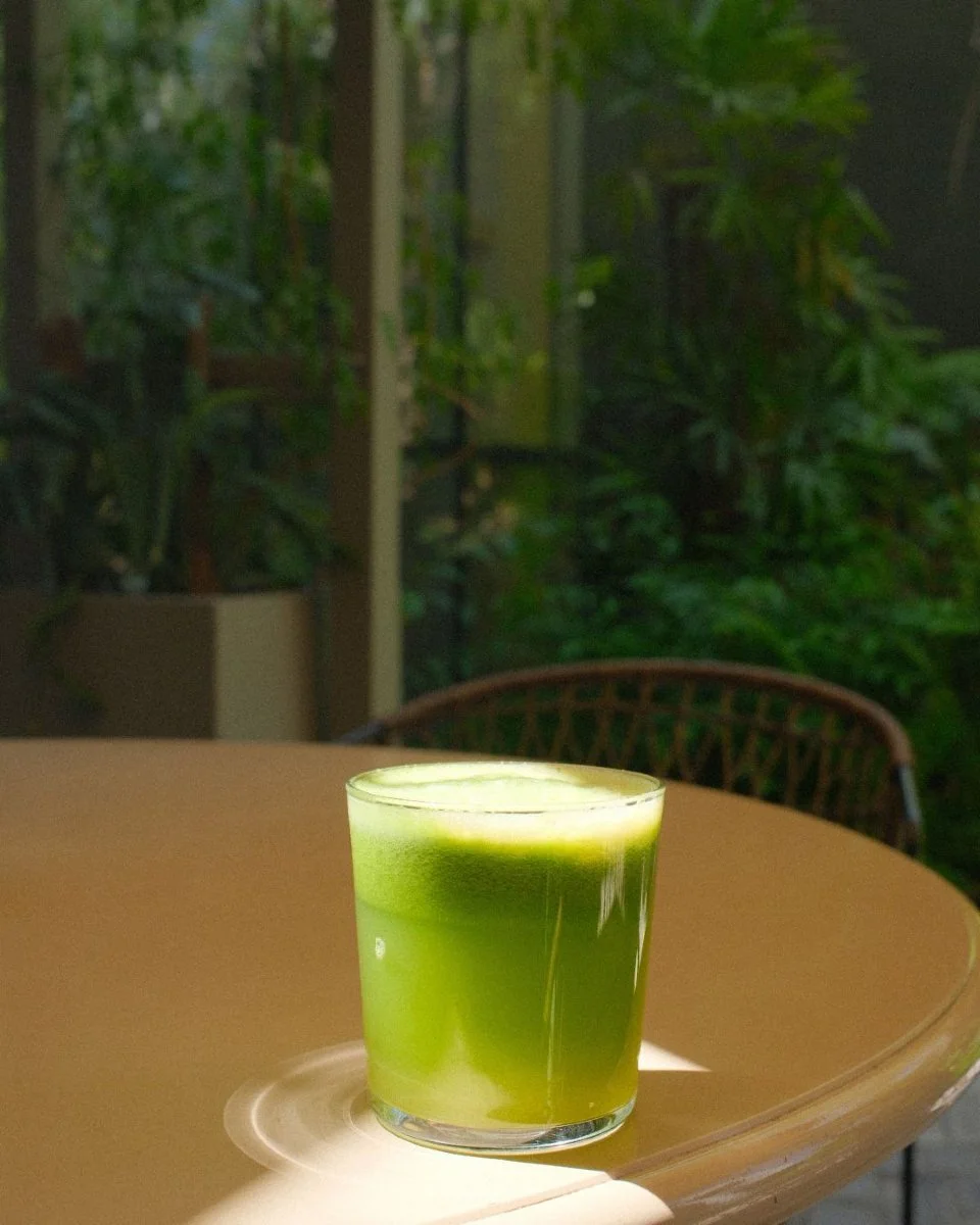 A clear glass of green juice on a table, with a blurred background of green plants and a brown chair.