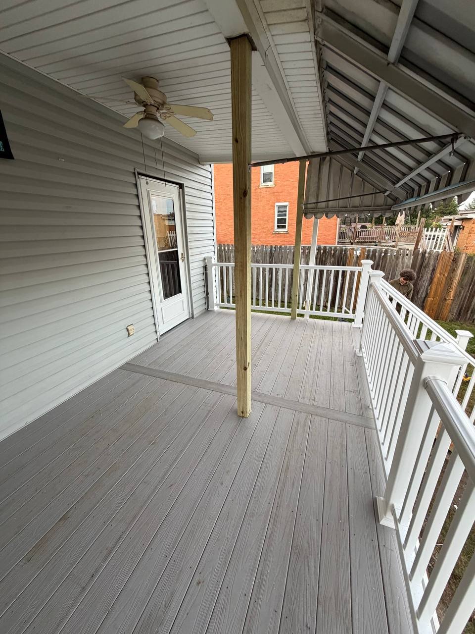 Back porch with gray wooden flooring, white railing, ceiling fan, and a door leading inside. Partially covered with an awning, overlooking a fenced backyard with a person near the fence.