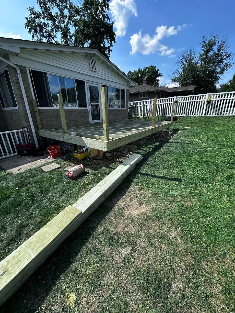 Construction of a wooden deck attached to the back of a house, with the framing in progress. The area shows construction tools and materials, with a grassy yard and a white fence nearby under a partly cloudy sky.