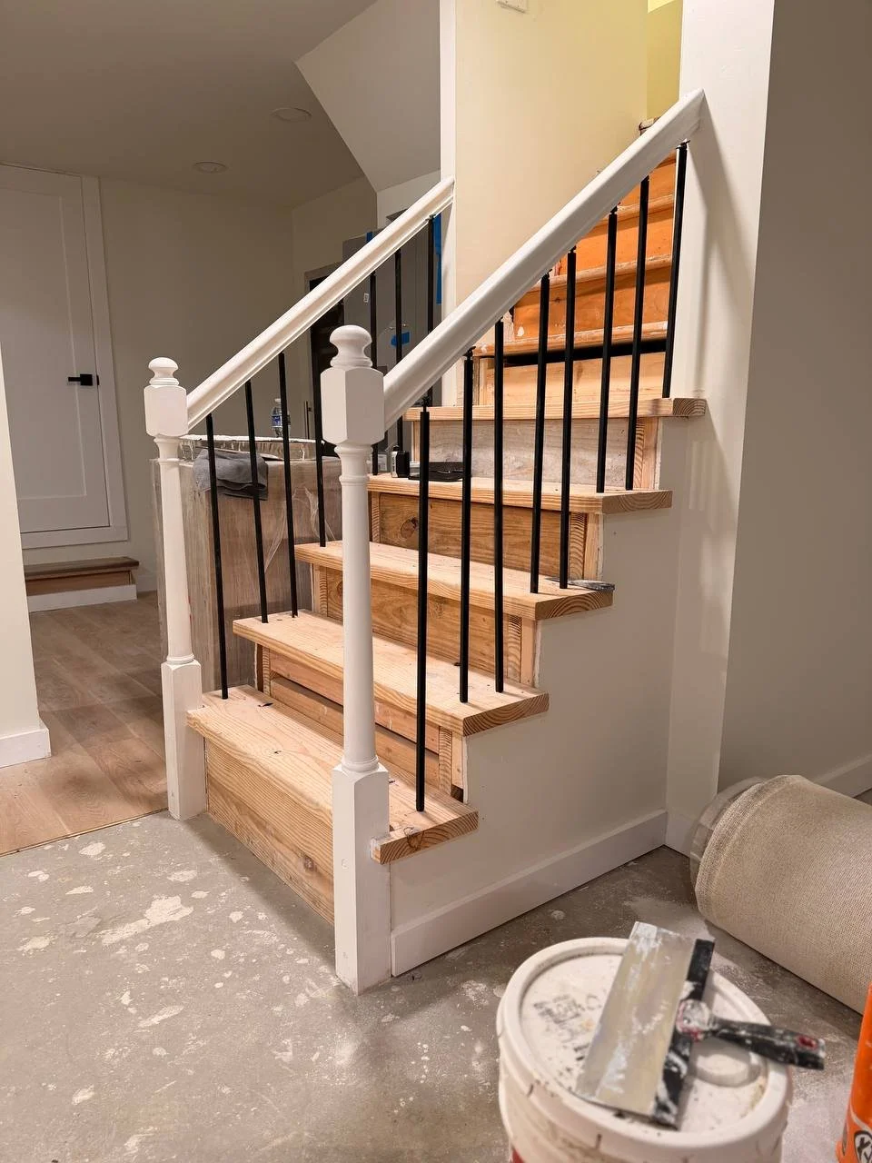 Interior of a house under renovation showing a partially built staircase with wooden steps, black metal spindles, and white painted newel posts. Construction tools and materials are visible on the floor.