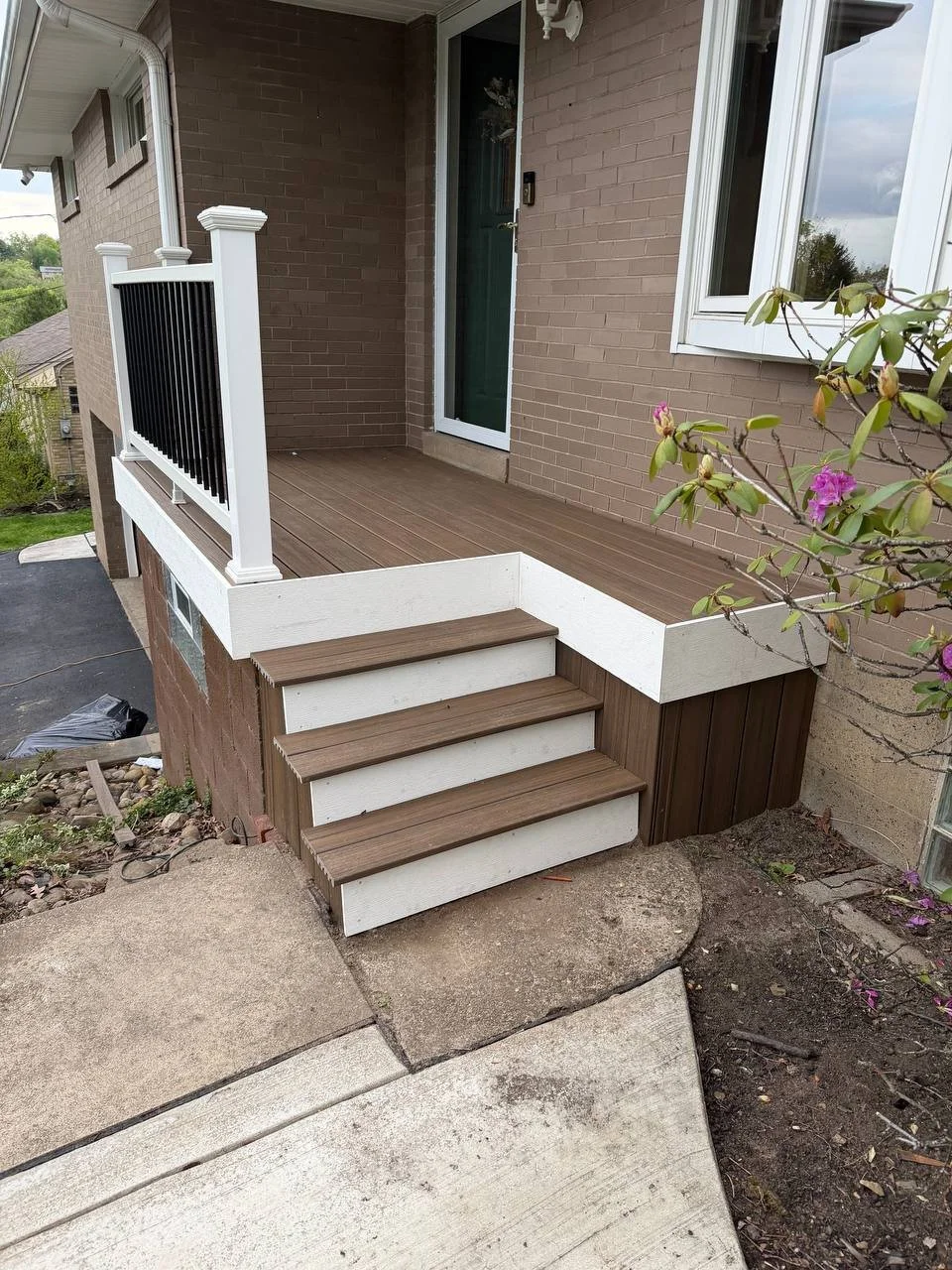 Small front porch with three steps leading up to a wooden deck, adjacent to a brick house. The porch has a black and white railing and a sliding glass door, with a window on the right side. There are plants with pink flowers near the porch.