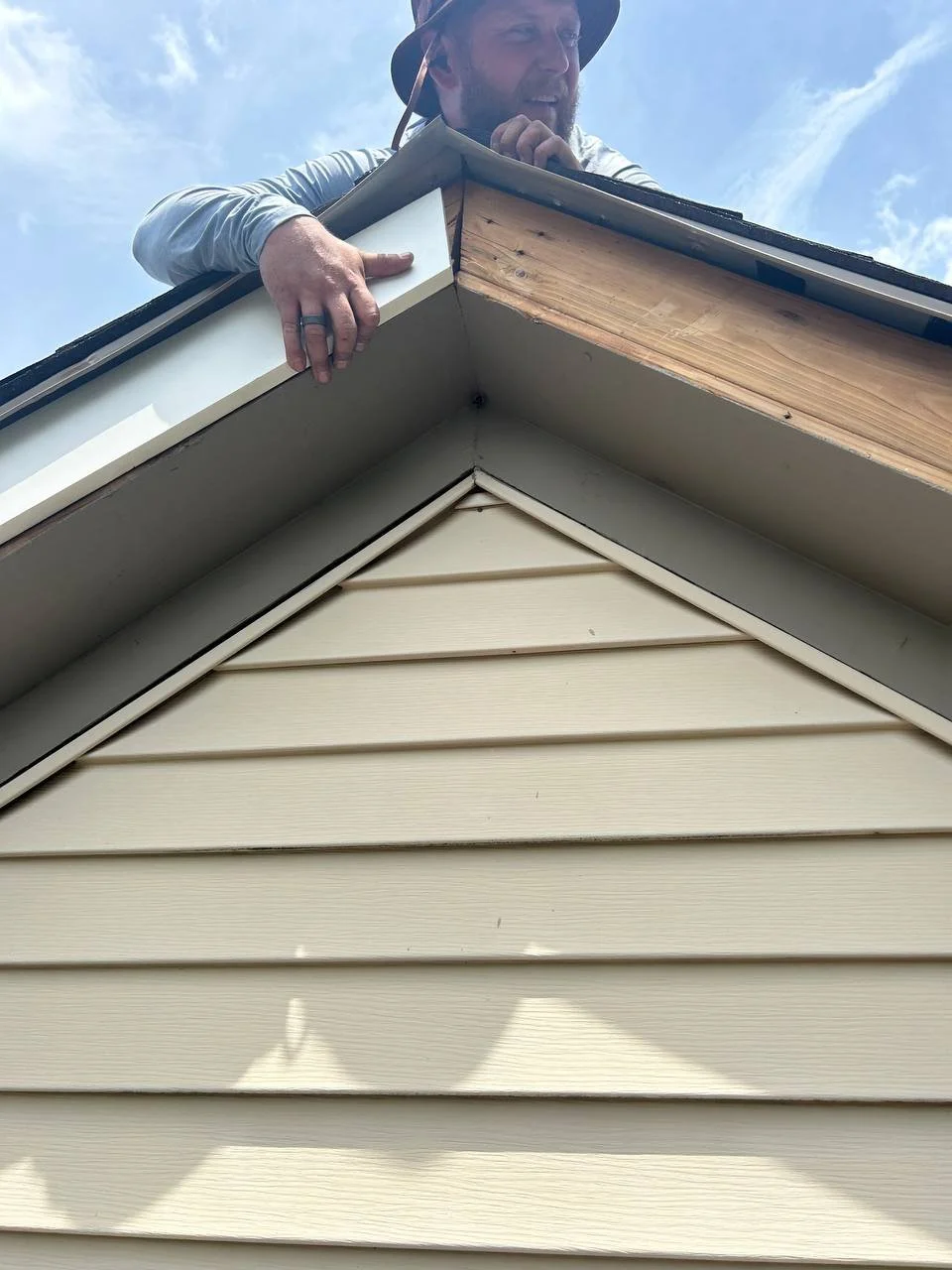 Man inspecting the roof of a house with beige vinyl siding, under a partly cloudy sky.