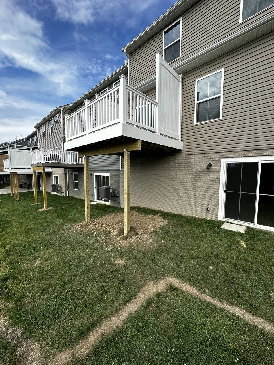 Multiple townhouses with decks, some still under construction, featuring white railings and support posts, with green grass and a partly cloudy sky.