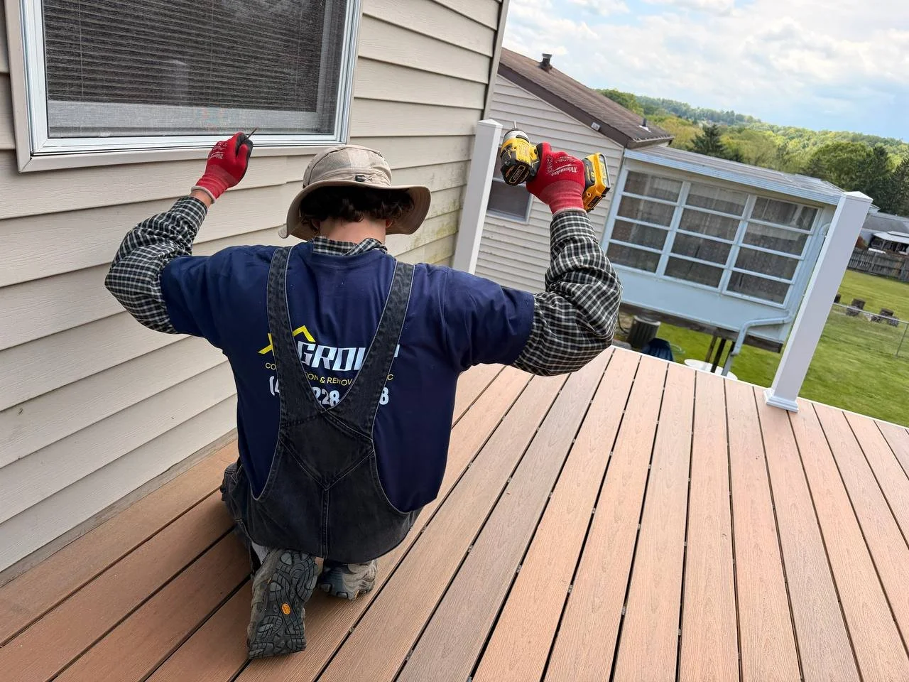 Person working on a wooden deck, holding a power drill in one hand and a screwdriver in the other, wearing gloves, a wide-brim hat, and a work shirt with a company logo.
