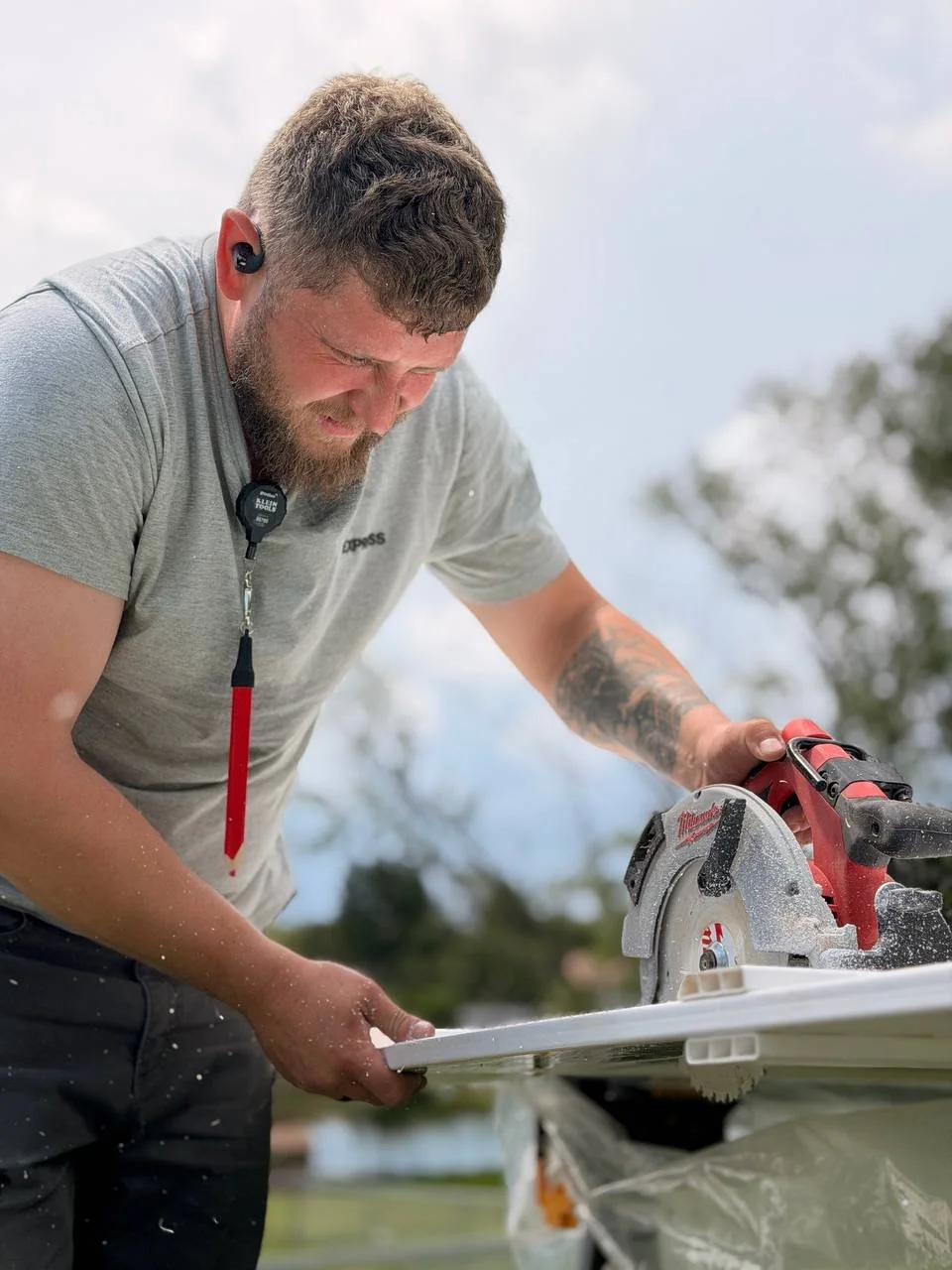 Man with beard cutting white wood using a circular saw outdoors, wearing a gray T-shirt and an earpiece.