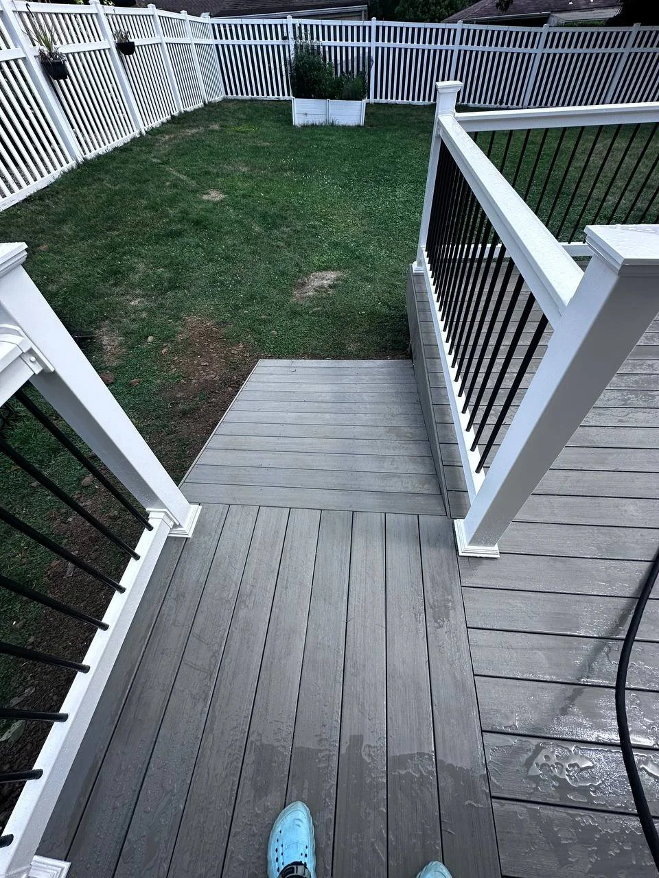 View from a deck with gray composite decking, looking out over a small grassy backyard enclosed with white picket and black railing fences. A person wearing white shoes stands on the deck.