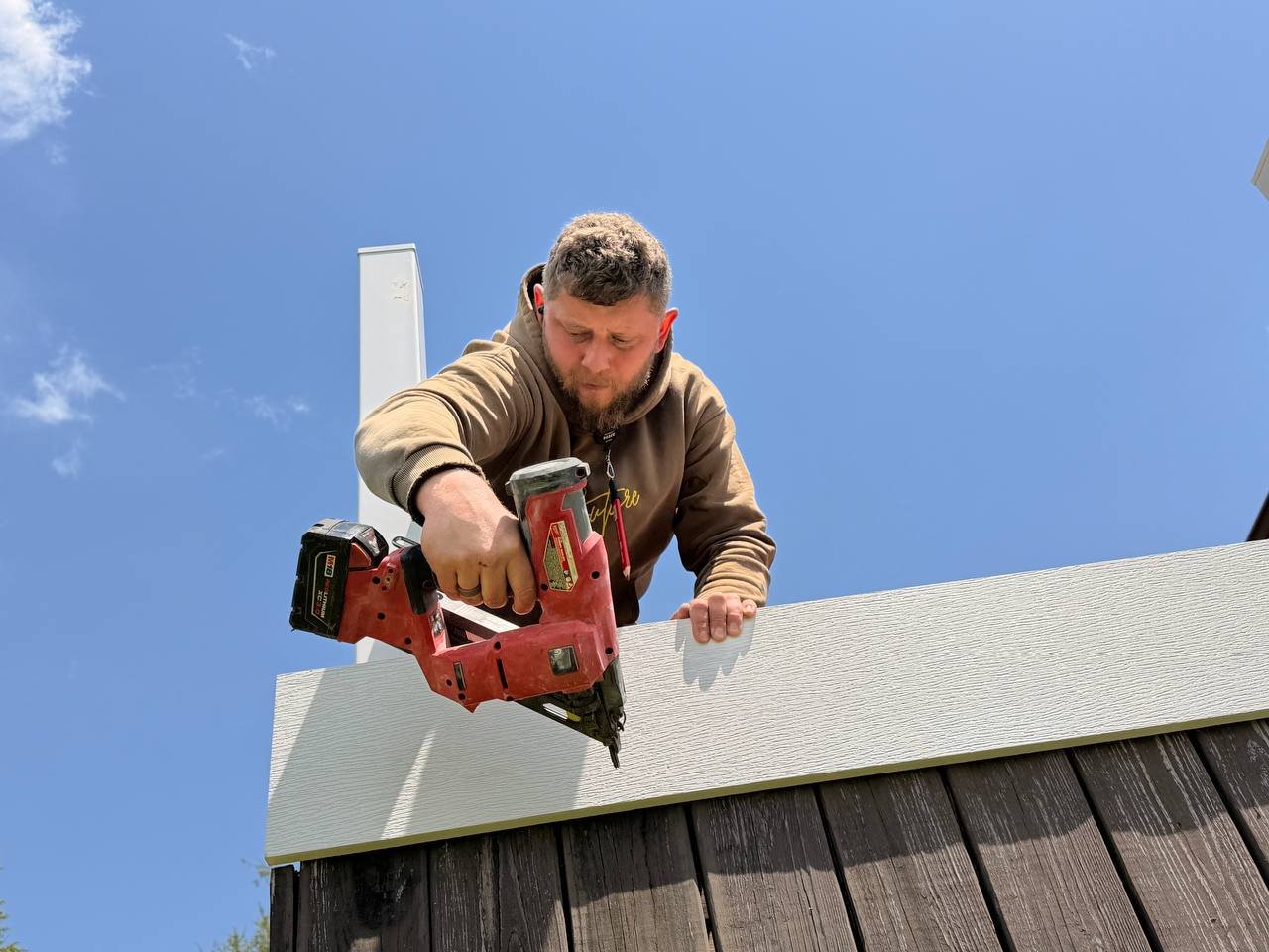 Man using a cordless nail gun to attach siding to a building under a blue sky.