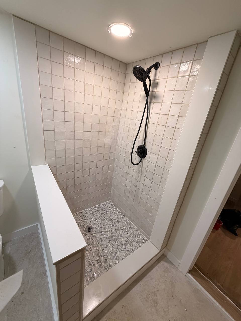 A tiled shower stall with a black showerhead and a wall-mounted control, surrounded by beige tiles with a small mosaic tile floor and a drain in the center.