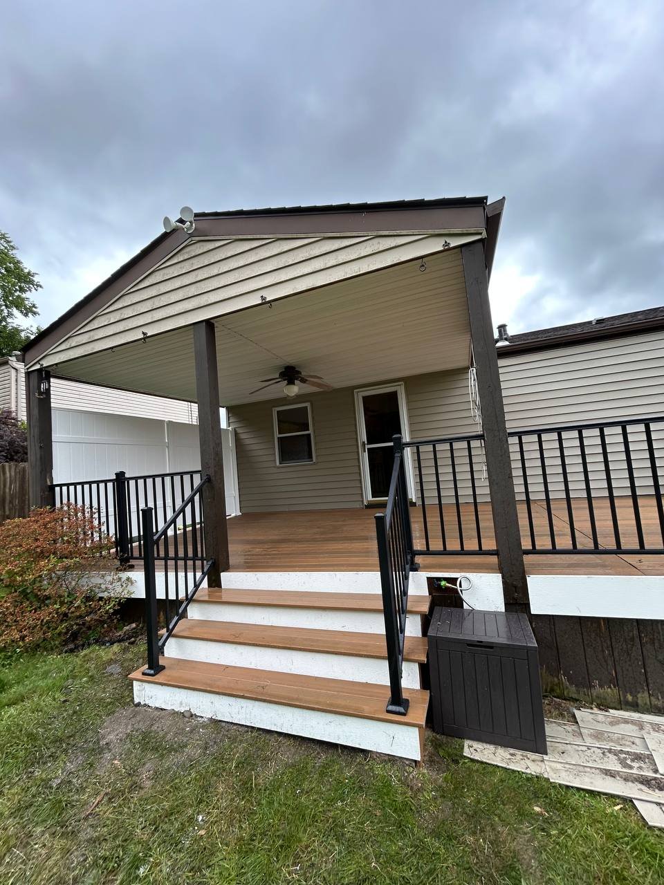 Newly built front porch with wooden flooring, black railings, and stairs leading to a grassy yard, with an overcast sky above.