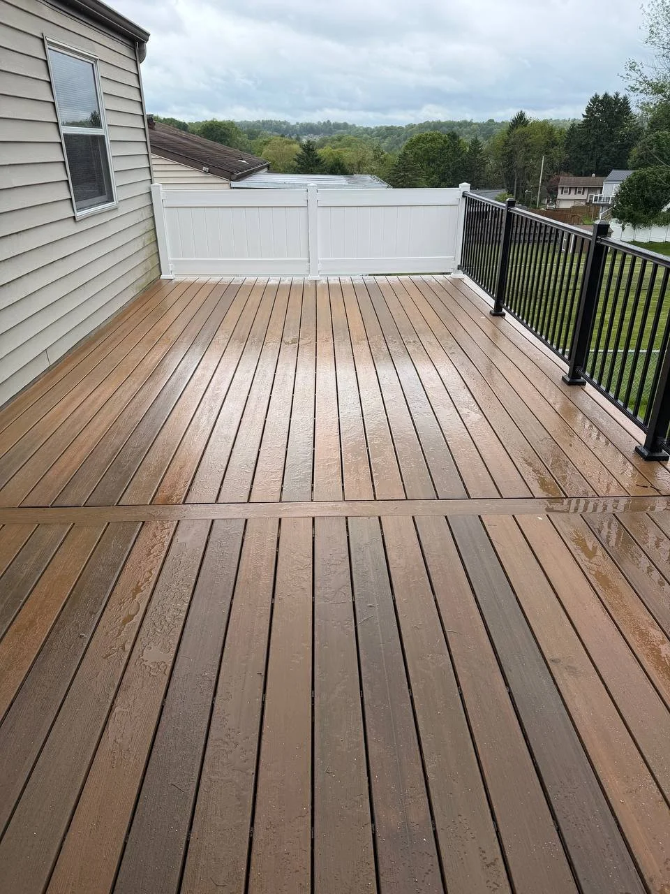 Wet wooden deck with black and white railings along the edge, overlooking a suburban neighborhood with trees and houses under a cloudy sky.
