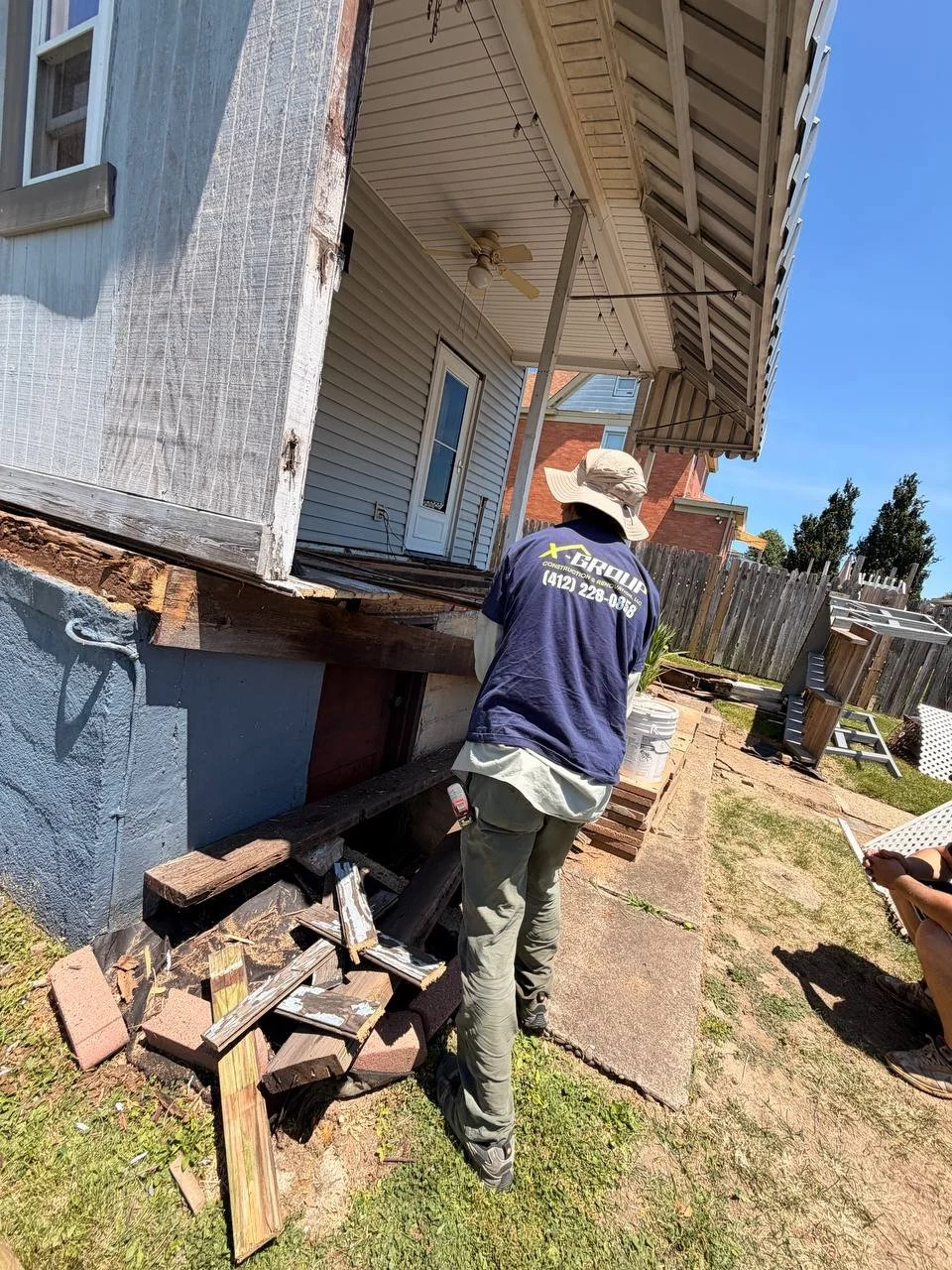 Construction worker inspecting the foundation of a house. The house has a balcony with a ceiling fan and light. Debris and bricks are scattered on the ground, and there is a wooden fence in the background.