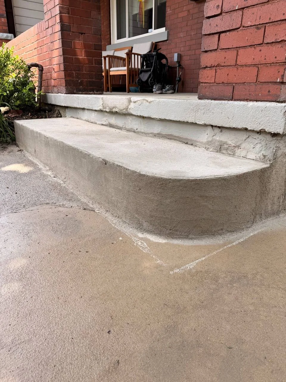 Concrete sidewalk and steps in front of a brick house with a window, outdoor chairs, and various items.