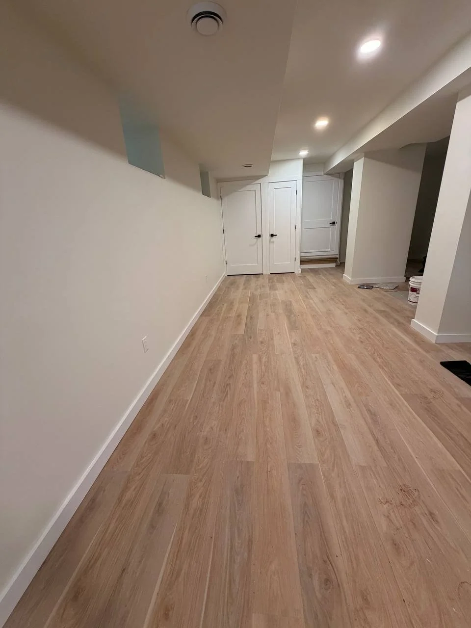 Empty room with light wood floors, white walls, and ceiling with recessed lighting. Closed white doors at the end of the room, with some construction tools and materials on the floor.