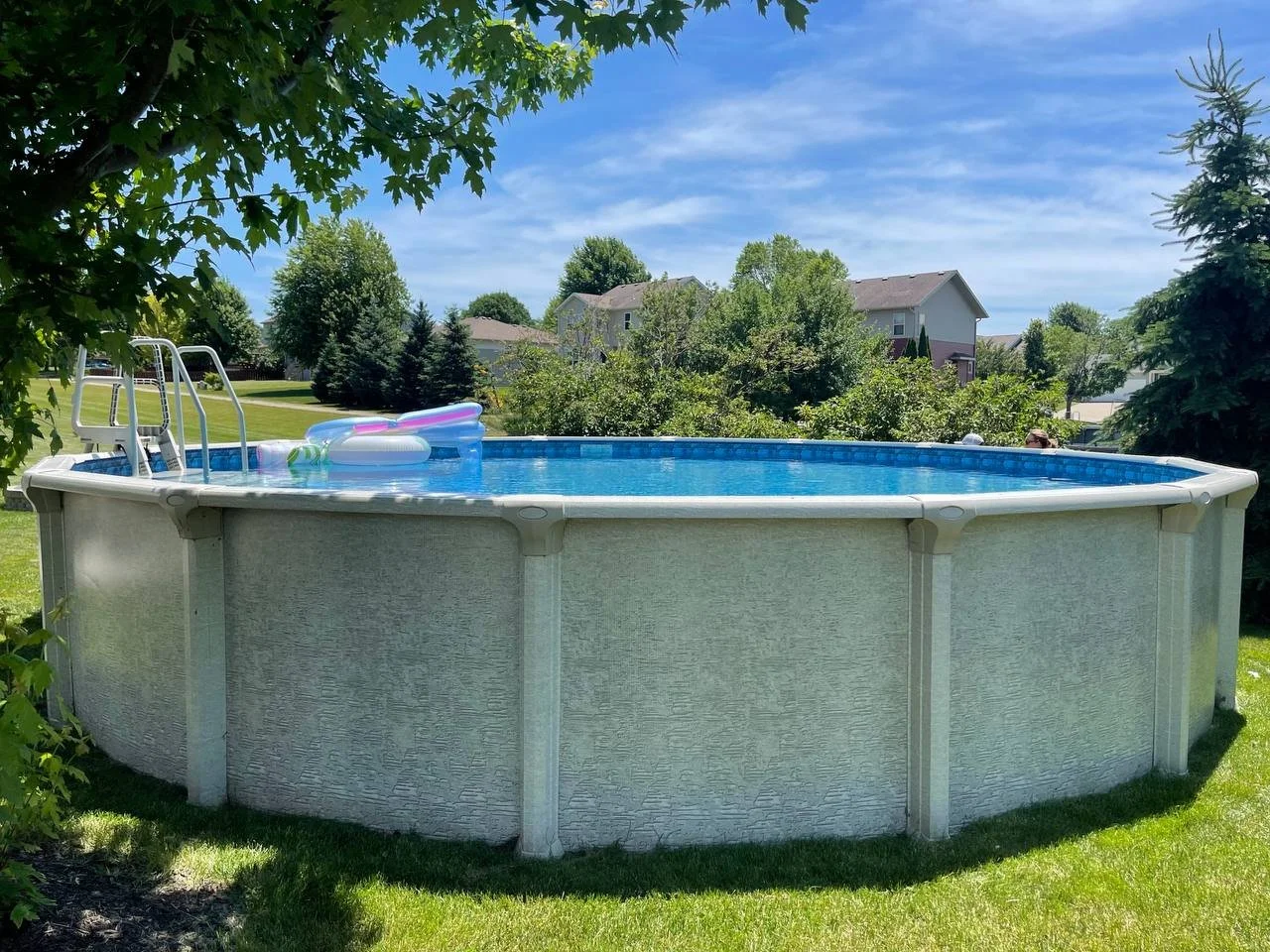 Above ground residential swimming pool in a grassy backyard with inflatable floaties inside, trees and houses in the background under a blue sky.