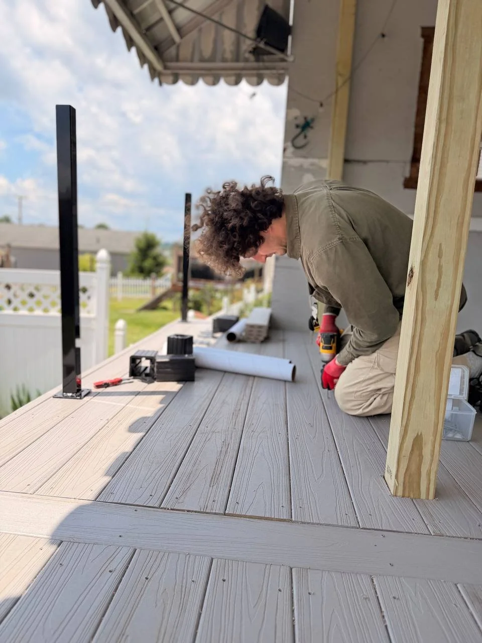 Person kneeling on a deck under construction, working on the railing or frame with tools and construction materials nearby outside a house.