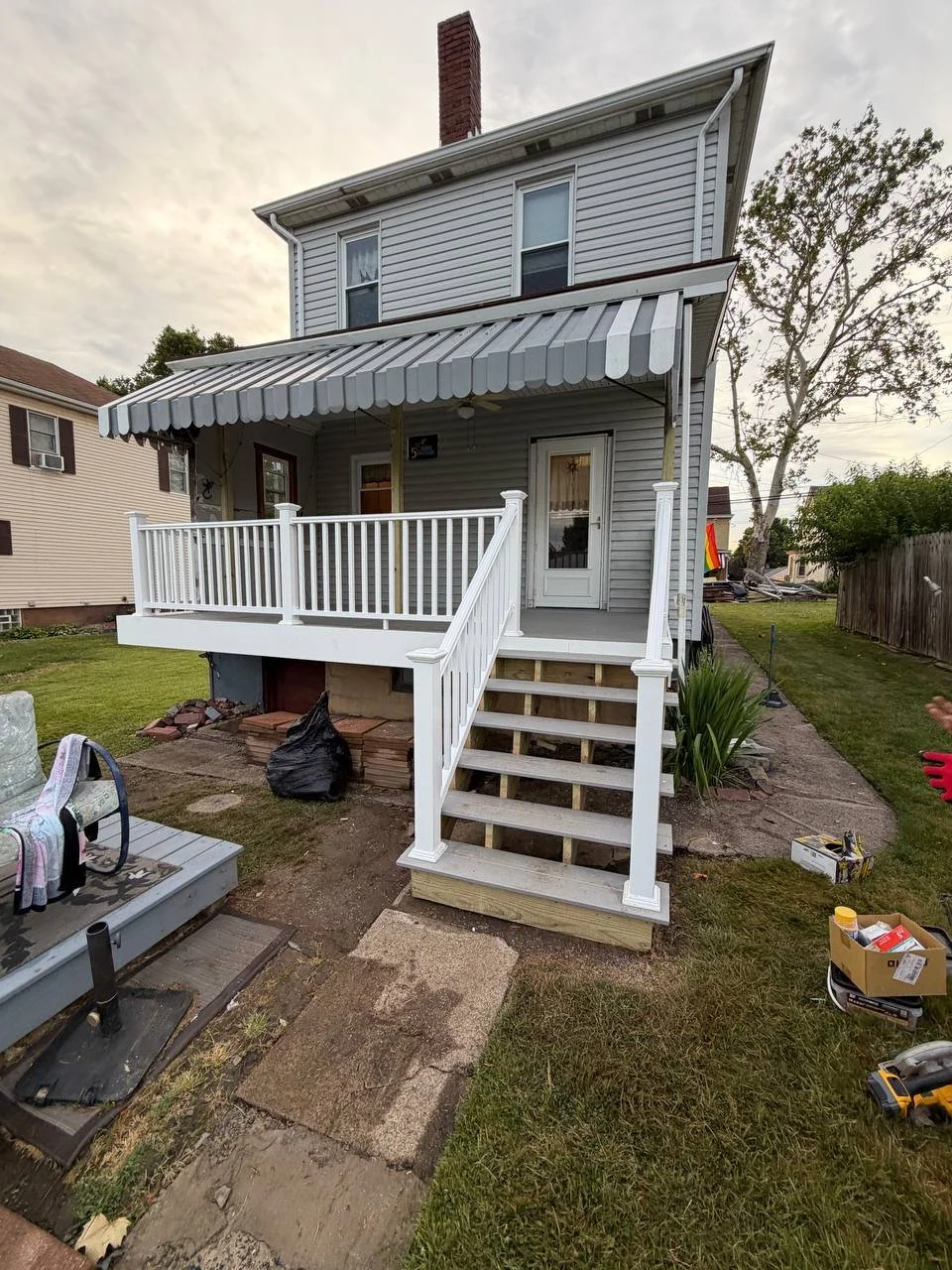 Two-story house with a small porch, white railing, and a striped awning over the door, showing ongoing exterior renovation with tools and materials around.