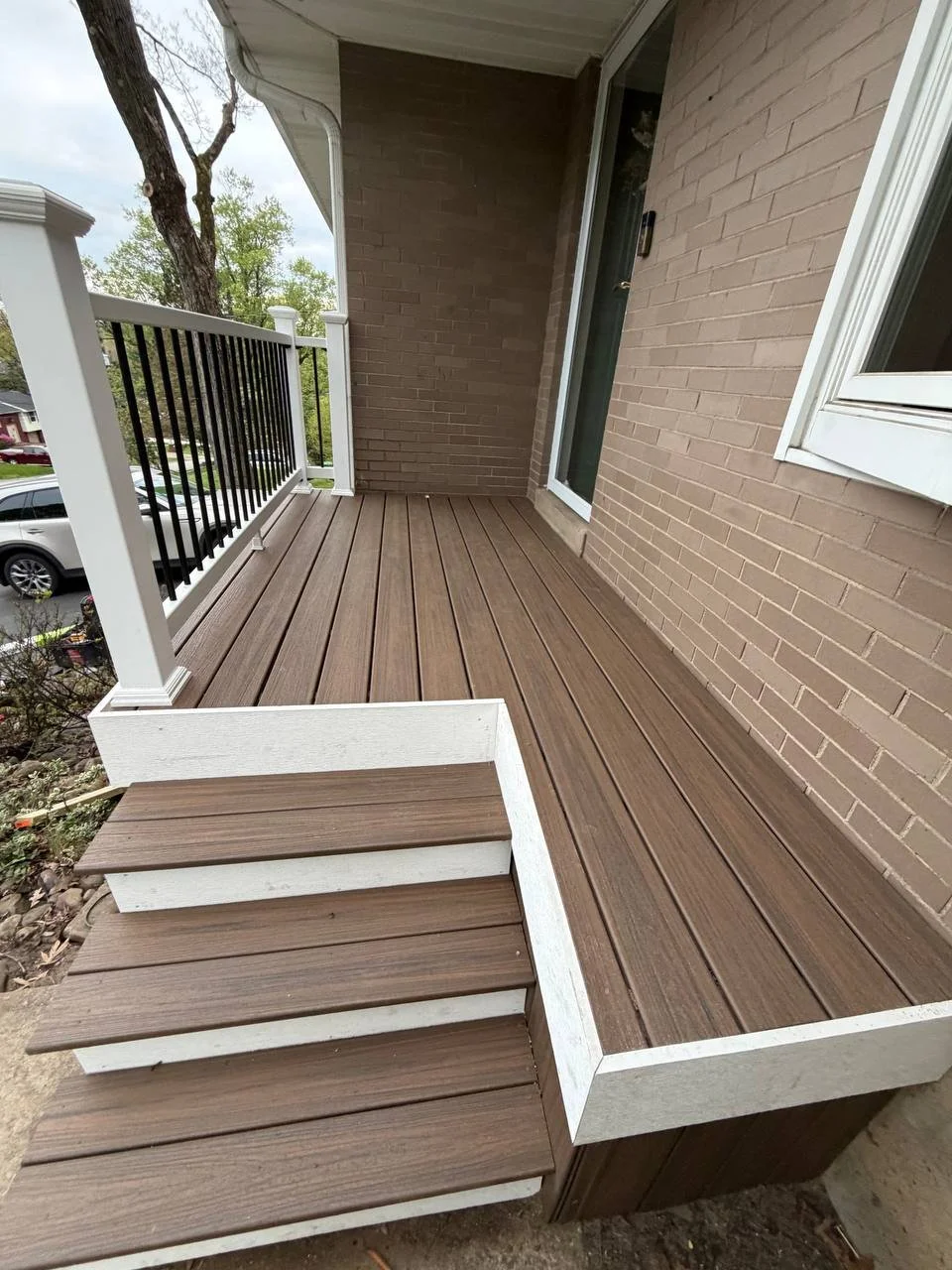 Small wooden porch with steps leading down to ground, attached to a brick house with a sliding glass door and a window, overlooking a residential neighborhood with trees and parked cars.