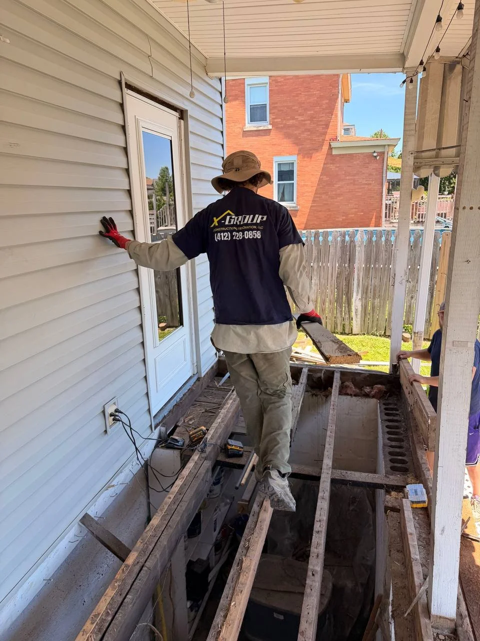 A construction worker standing on the edge of a house's porch framing, wearing a hat, gloves, and work clothes, working near a door while two children observe nearby.