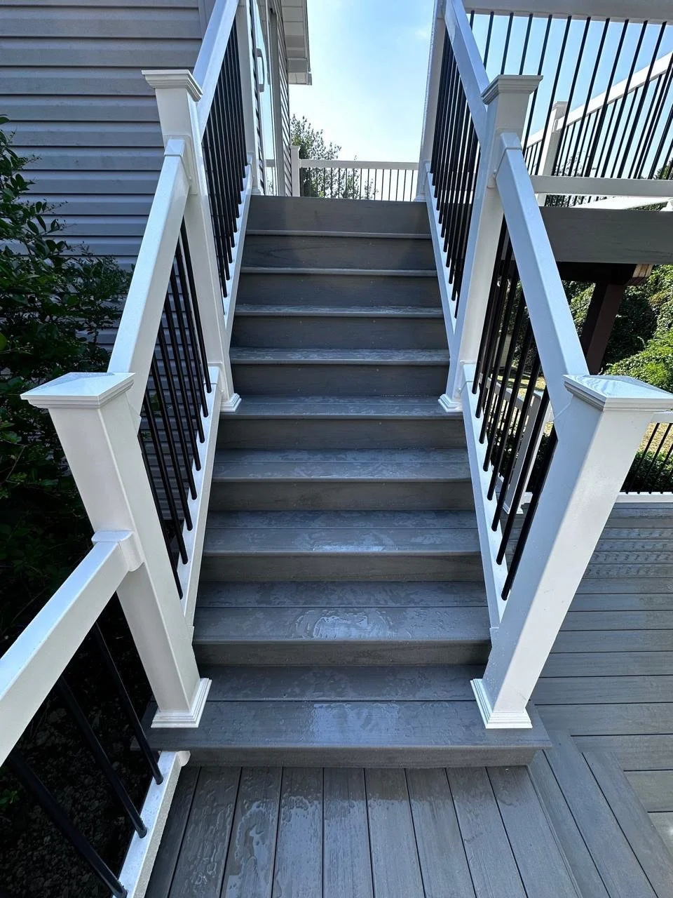 An outdoor staircase with gray wooden steps and white railings, leading to a second-story deck with black balusters, beside a house with white siding.