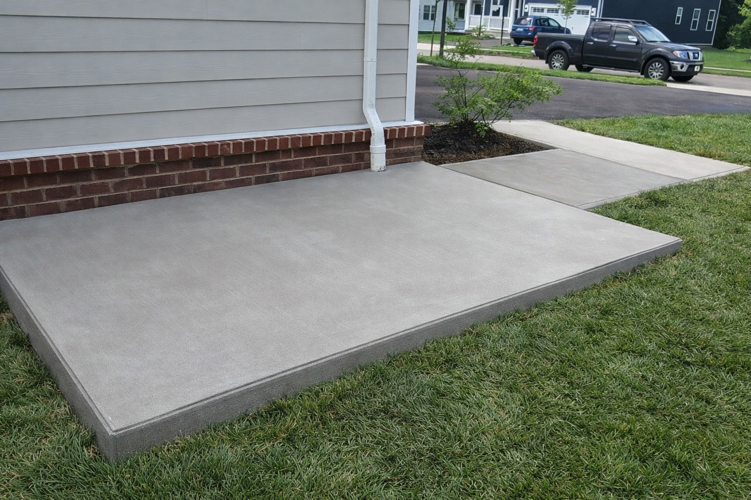 Concrete porch with brick skirting and an adjacent sidewalk in front of a house with vinyl siding, visible grass, and parked cars on the street.