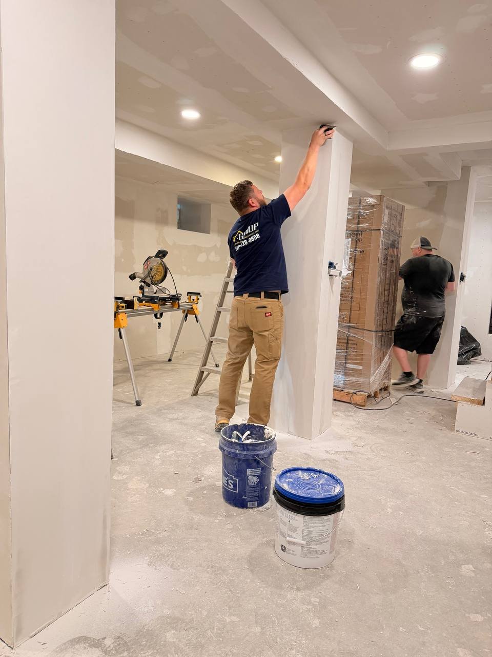 Two workers painting walls in a room under construction, with painting tools and buckets of paint on the floor.
