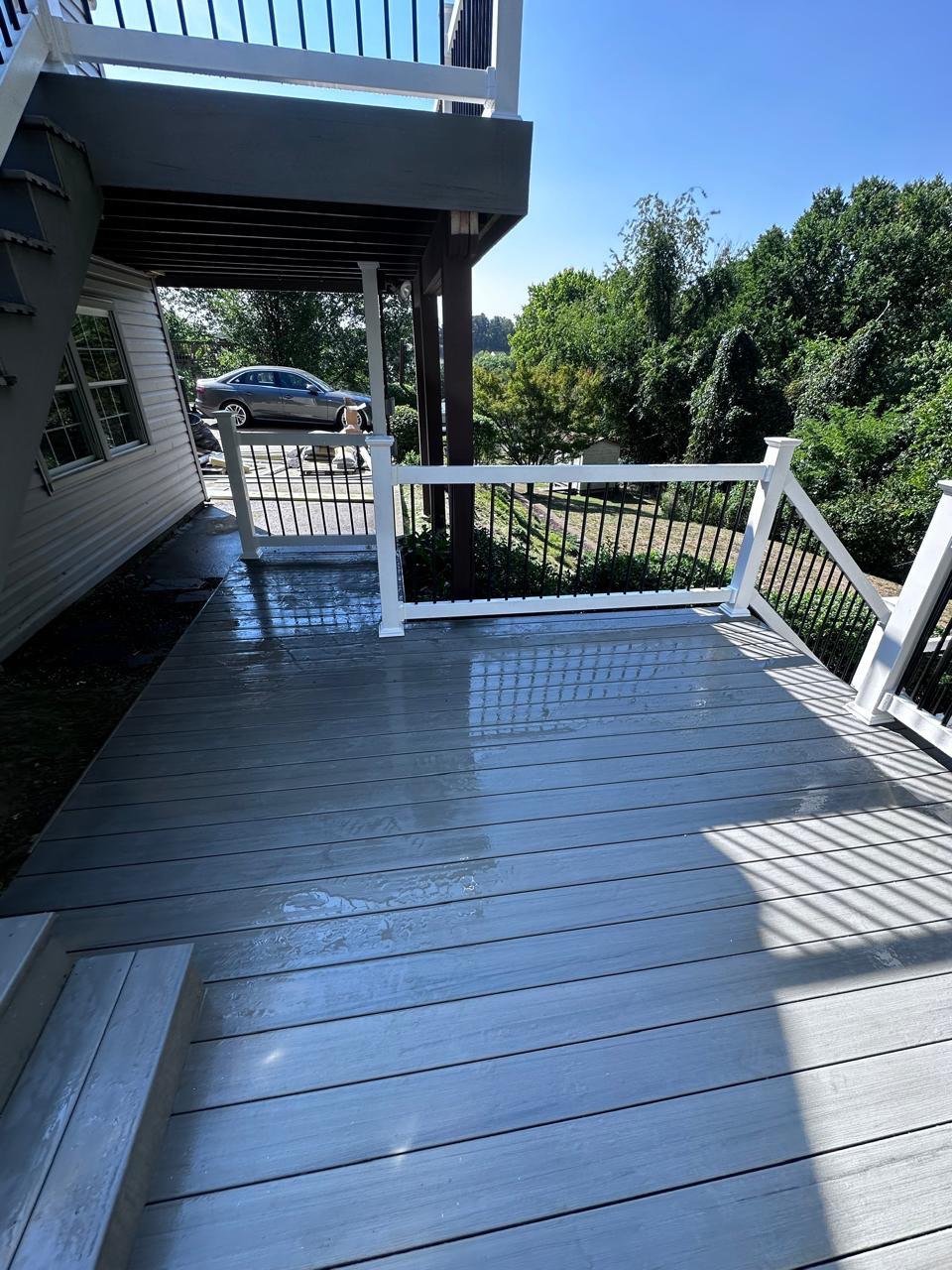 A freshly cleaned outdoor deck with wet surface and a white railing, overlooking a green backyard with trees and a driveway where a black car is parked.