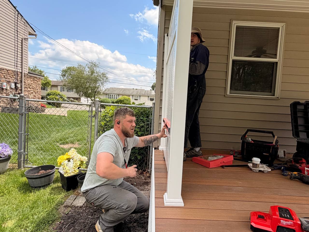 Two men installing a white pillar on a porch, with tools and a toolbox nearby. One man is squatting outside near flower pots, and the other is standing on the porch working on the pillar.