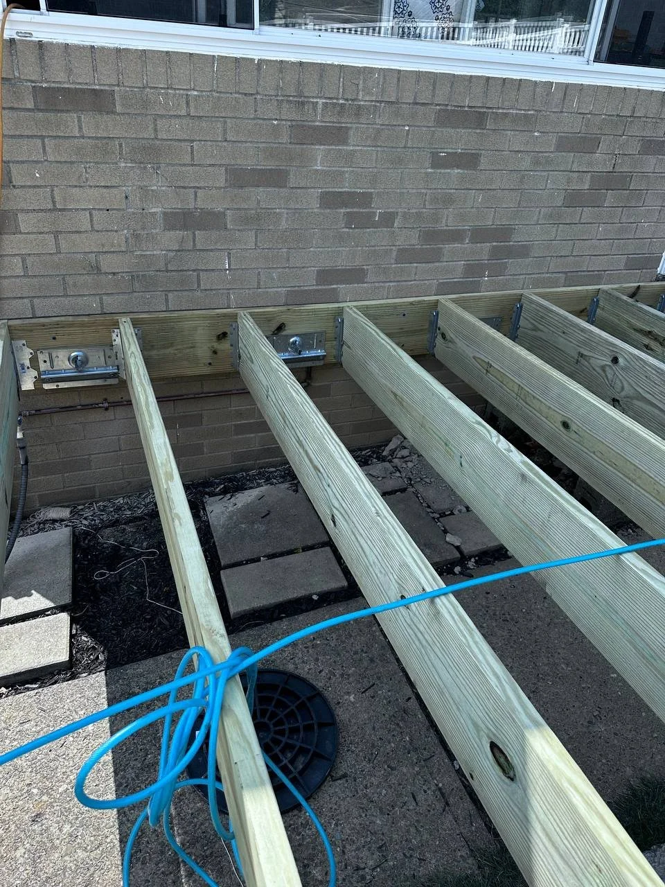 Wooden deck framework under construction against a brick wall, with blue electrical cord and concrete pavers on the ground.