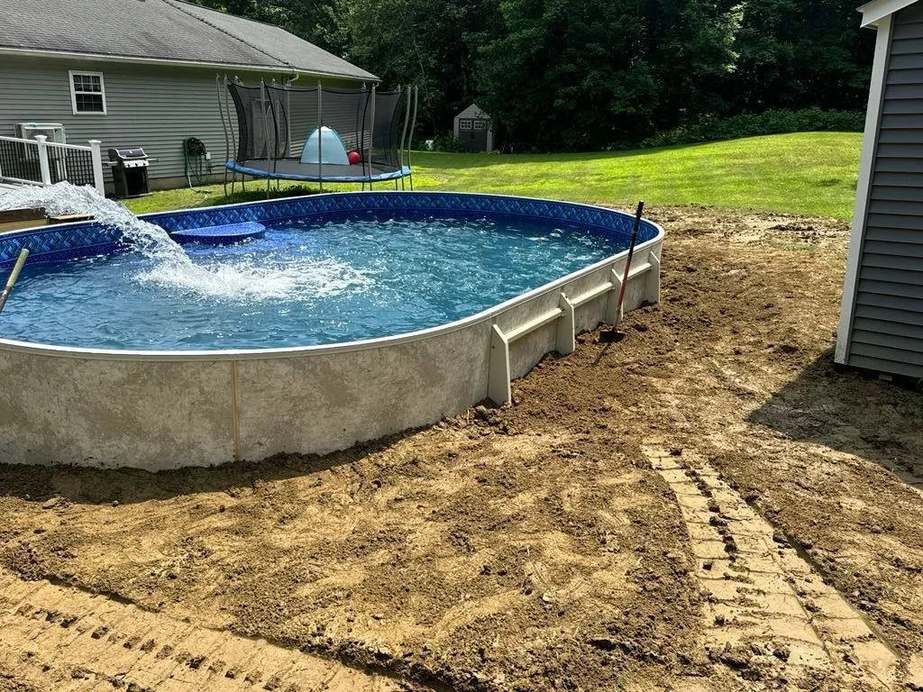 Above-ground swimming pool filled with water, situated on bare dirt with a path made of bricks leading to it. A pool skimmer is resting on the pool's edge, and a pole with a net is submerged in the water. In the background, there is a trampoline with