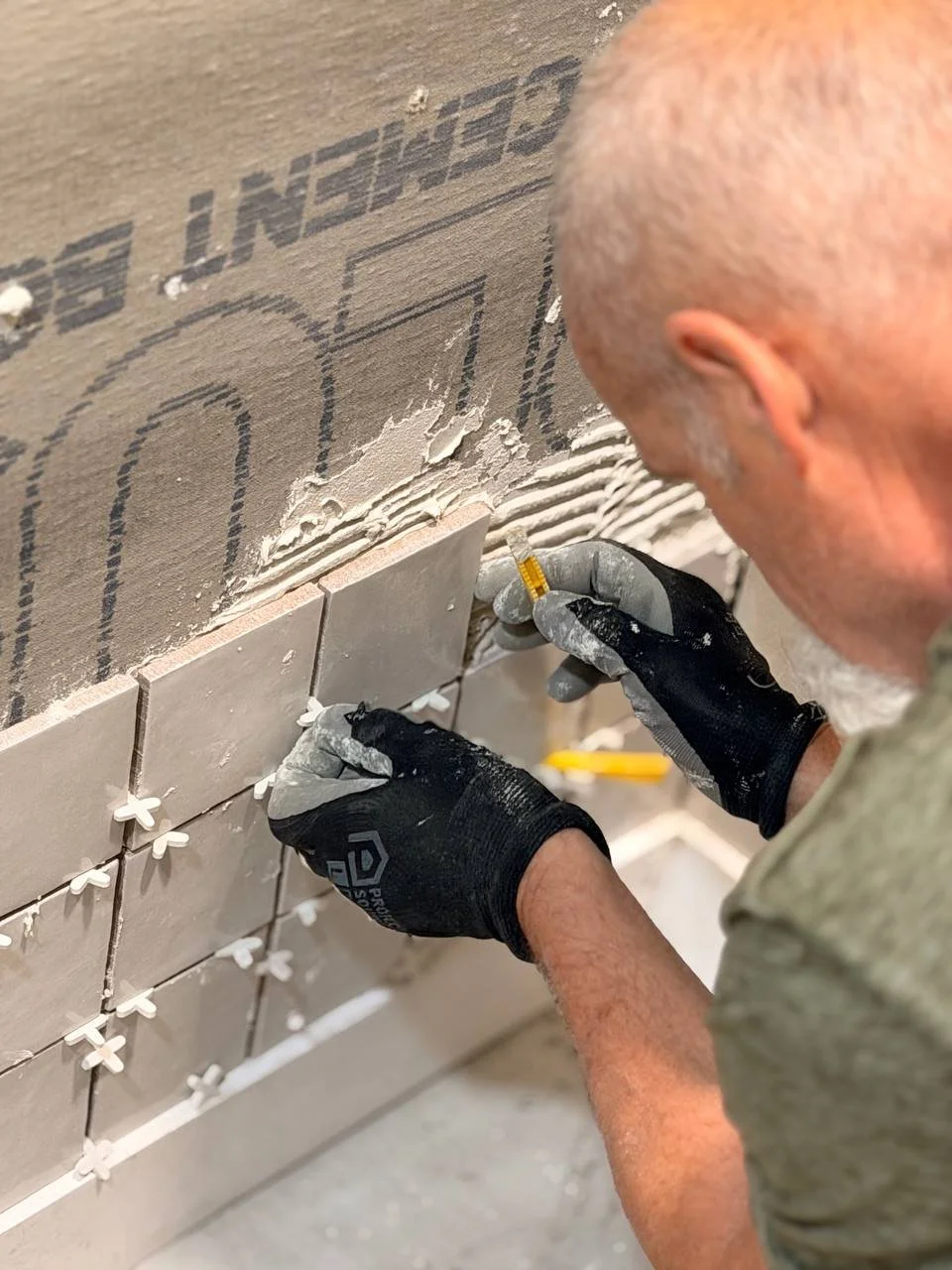 A person wearing black gloves is installing tiles on a wall, using a level to ensure proper alignment, with drywall and marking lines visible in the background.