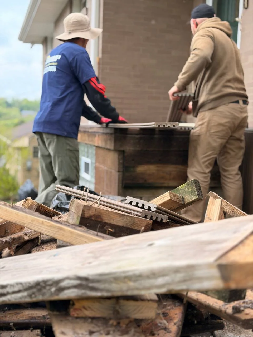 Two men working on a construction or renovation project outside a house, handling wooden planks and tools, with a pile of wood and construction supplies in the foreground.