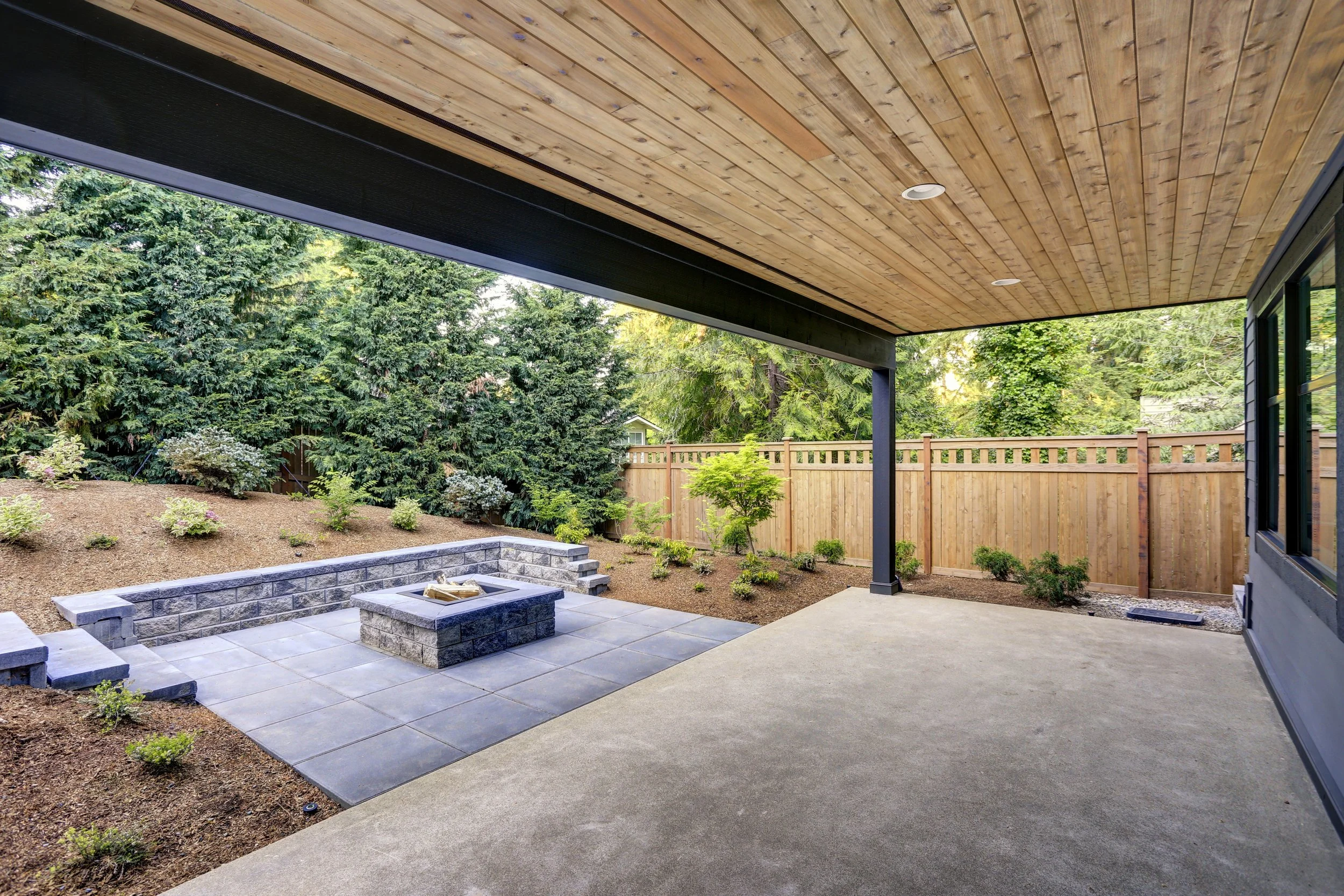 Backyard patio with a concrete patio, stone fire pit, and a landscaped yard with trees and bushes, enclosed by a wooden fence.