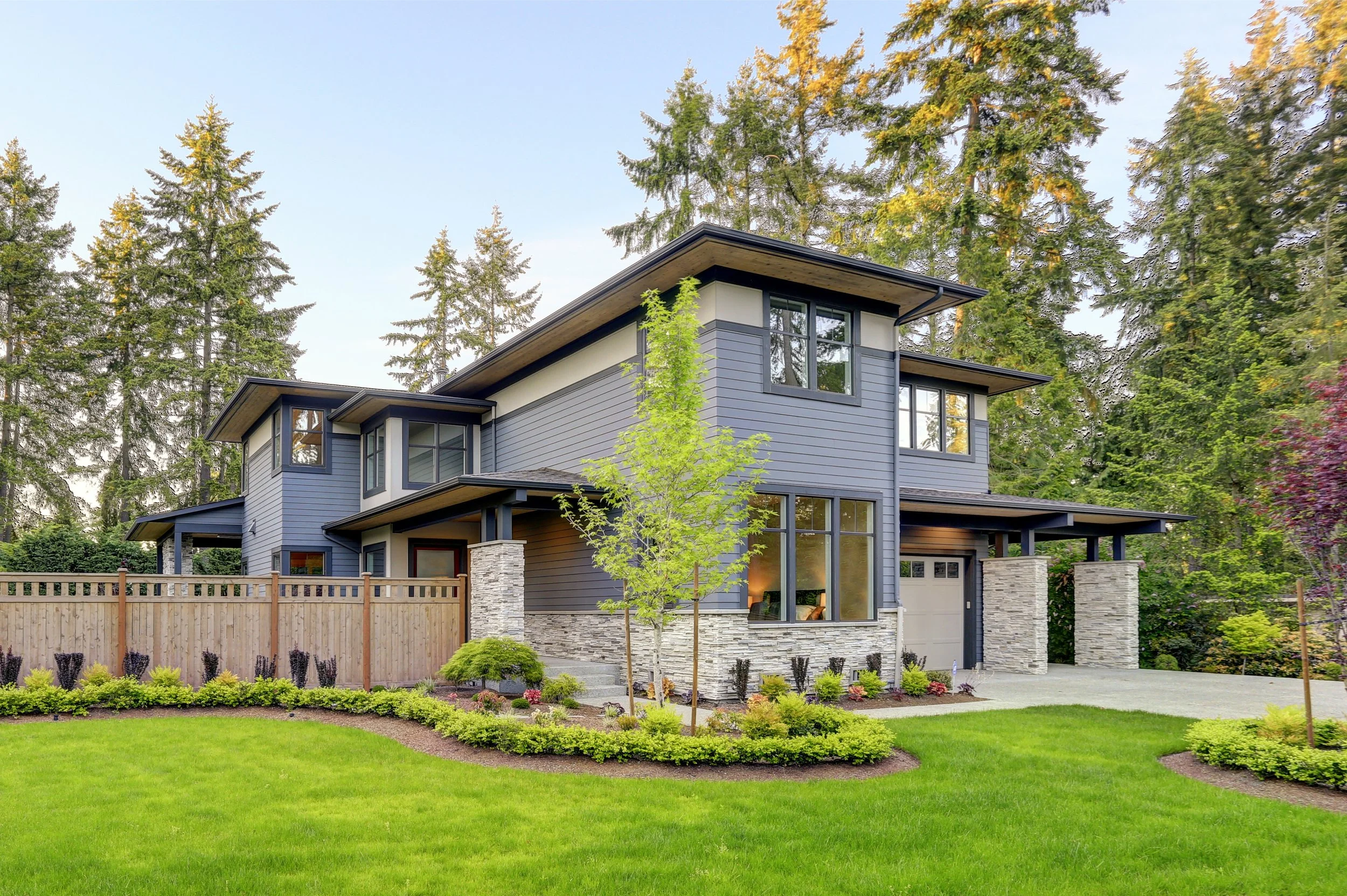 Modern two-story house with gray siding, stone accents, large windows, surrounded by a well-manicured lawn, bushes, and tall trees in the background.