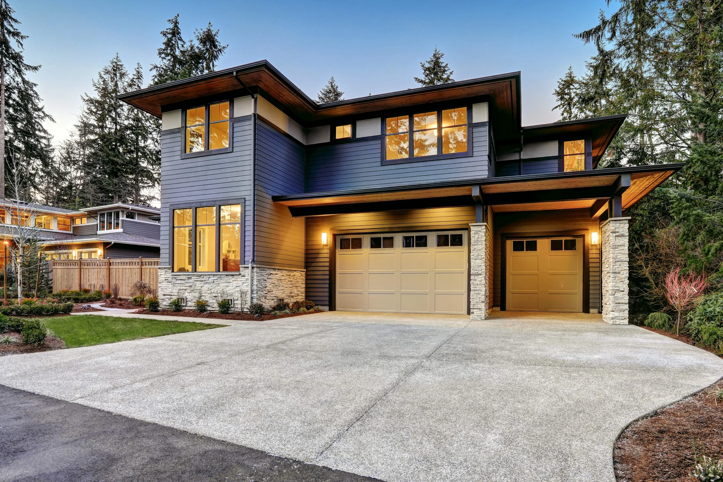 Modern two-story house with dark blue siding, stone accents, and large windows, surrounded by trees and a landscaped yard.