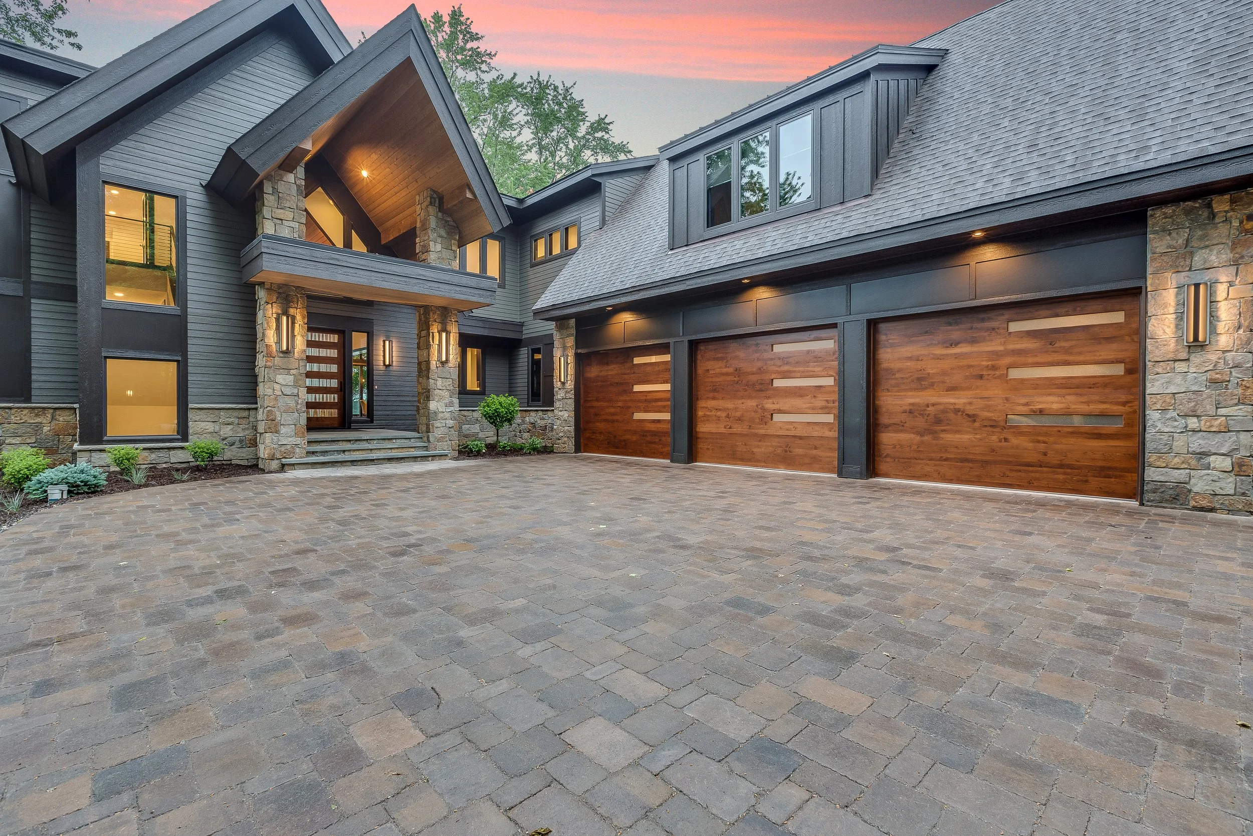Front view of a modern house with a driveway, stone and wood exterior, multiple garage doors, and a porch with stairs, during sunset.