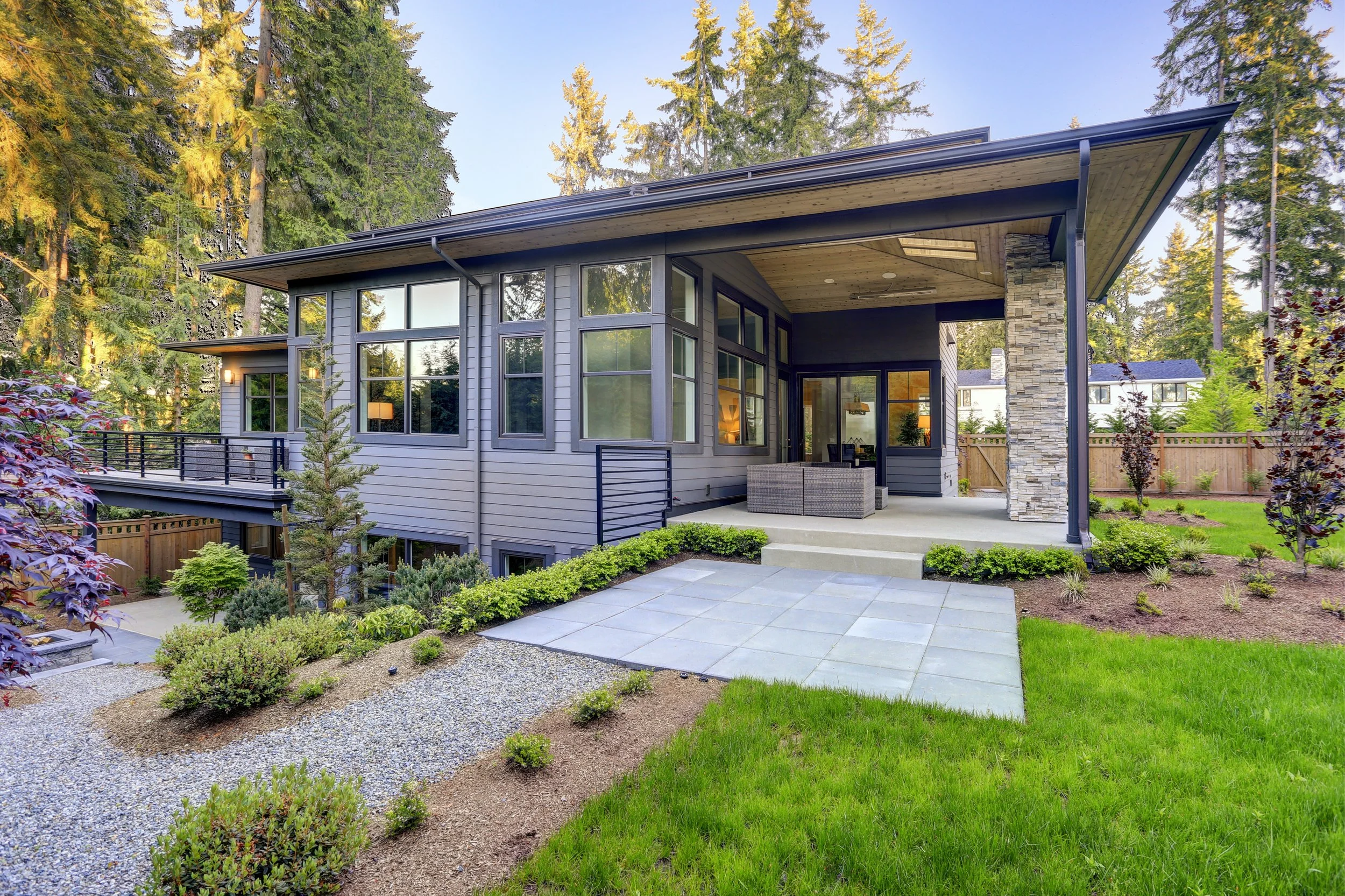 Modern house with large windows, a covered porch, outdoor seating area, and landscaped yard with grass, bushes, and small trees.
