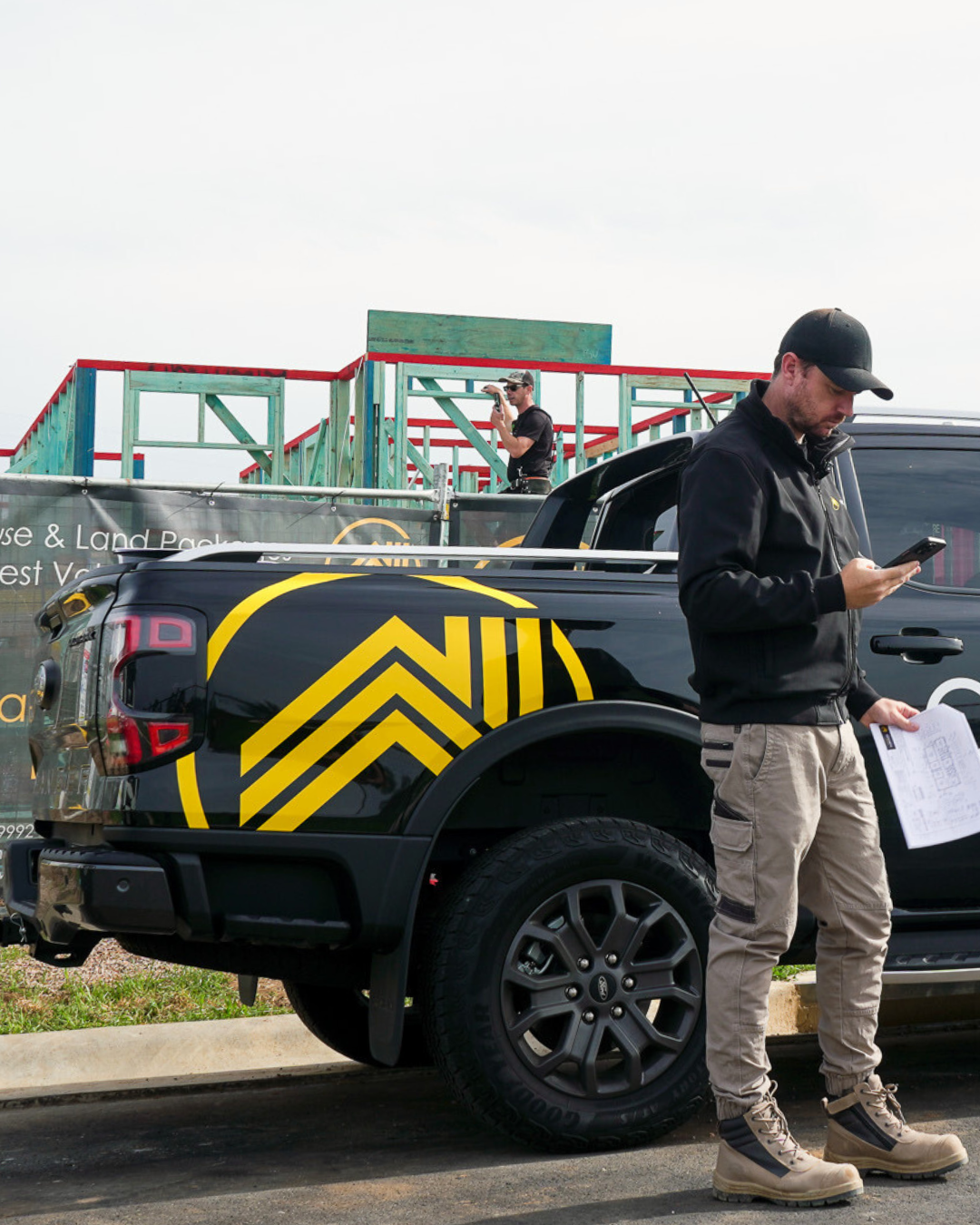 Two men near a black pickup truck with yellow and black graphics, working at a construction site with a partially built wooden structure in the background. One man is standing next to the truck looking at his phone, and the other man is standing on the truck bed, also using his phone.