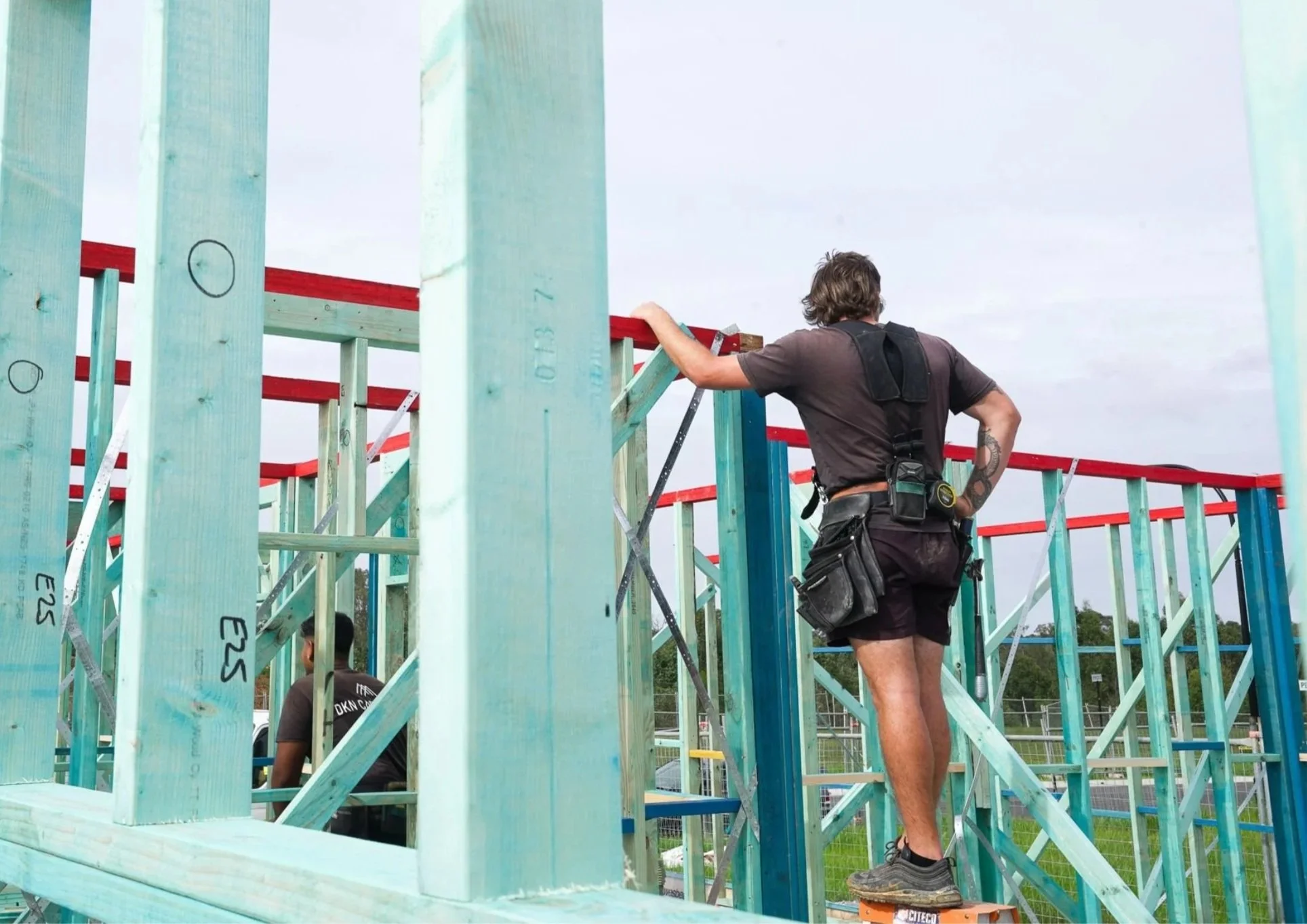 A construction worker standing on a small platform, working on a wooden building frame at a construction site outdoors.