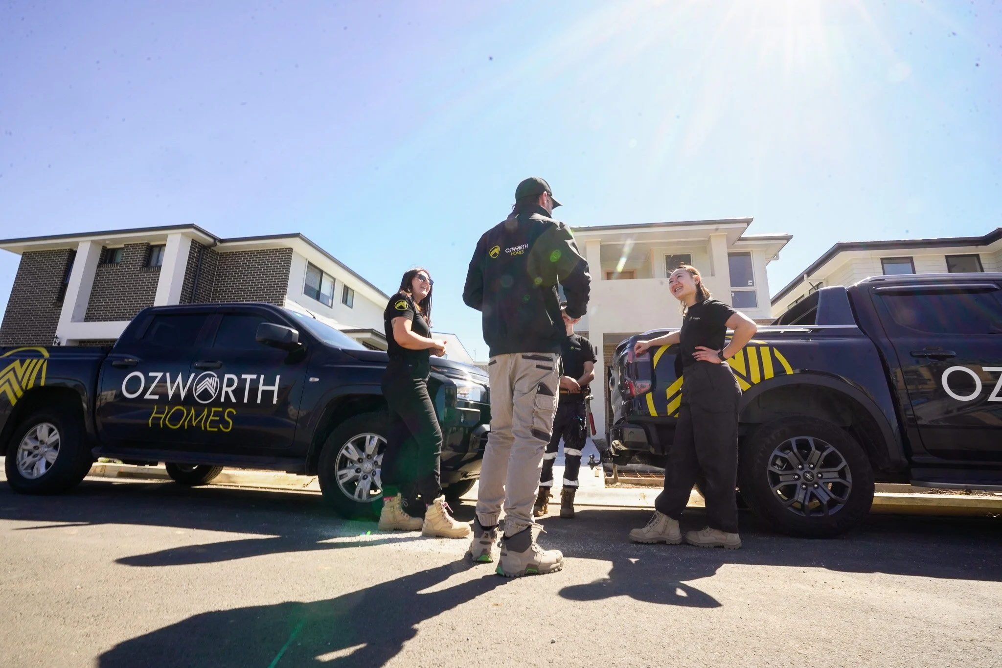 A group of people, likely real estate agents or construction workers, standing beside two Ozworth Homes trucks in a residential neighborhood, engaged in conversation on a sunny day.