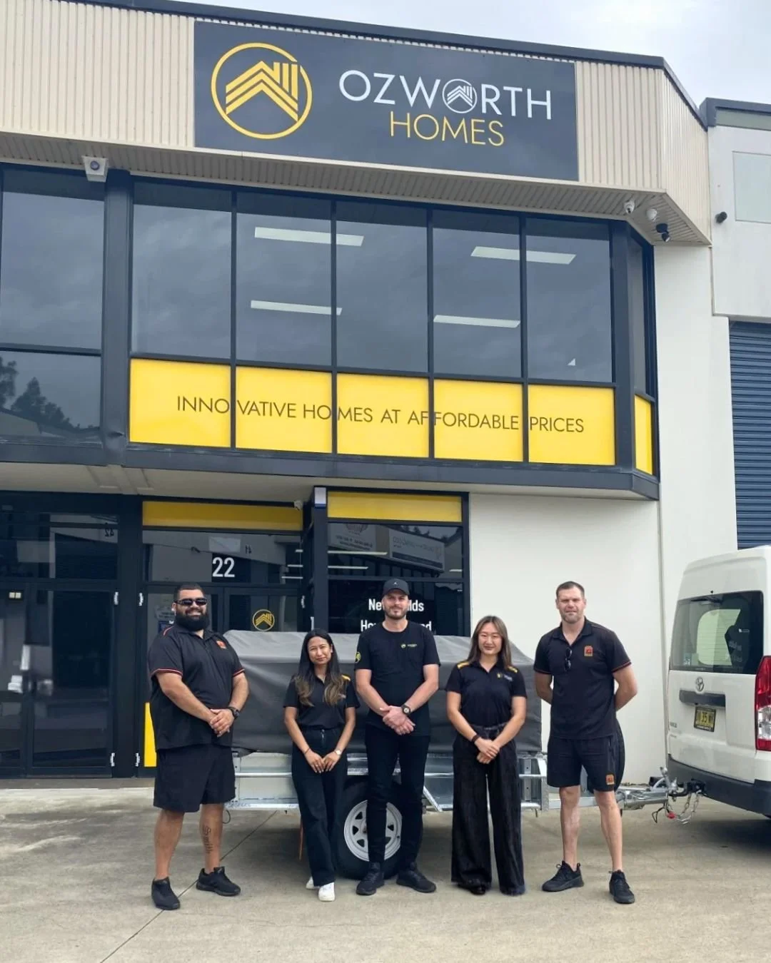 Group of five people standing in front of Ozworth Homes building, with a banner that reads 'Innovative Homes at Affordable Prices' and a trailer. The building has a sign with a house logo and the company name.