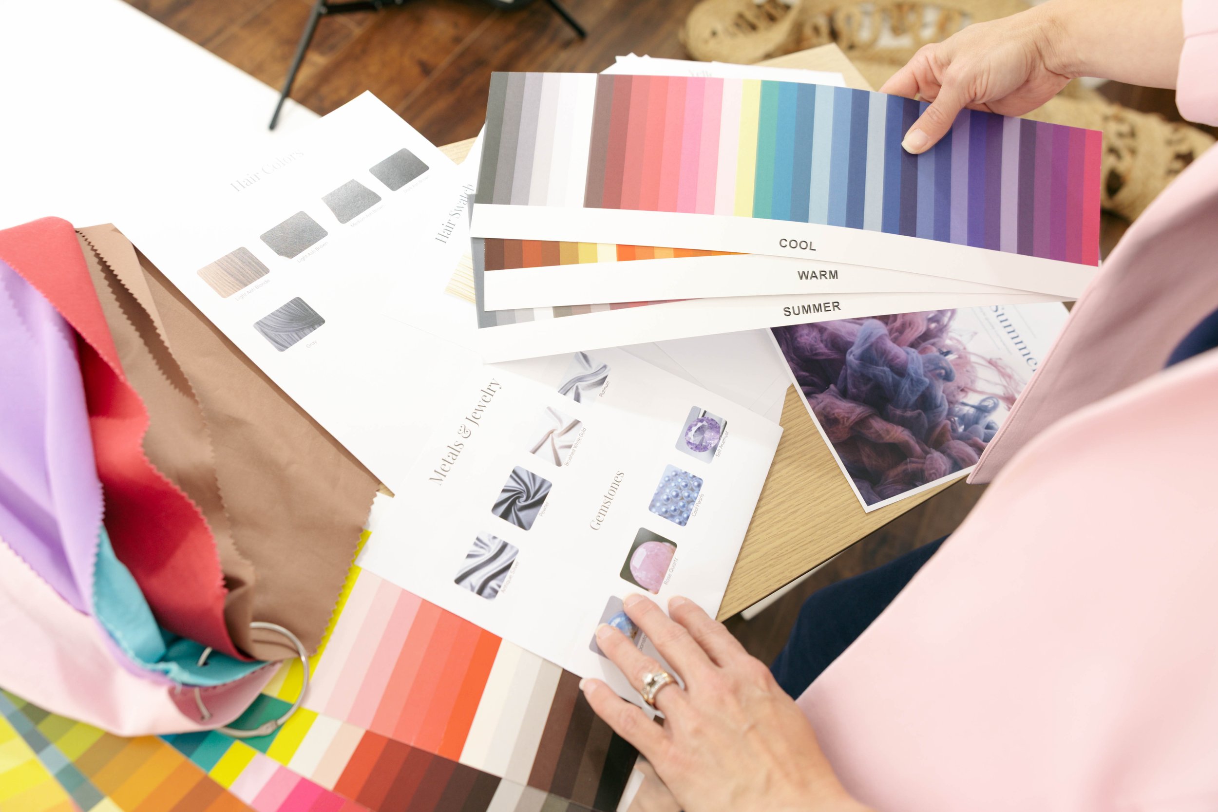 Person holding color swatches labeled 'COOL,' 'WARM,' and 'SUMMER' amidst fabrics and printed material on a wooden table.